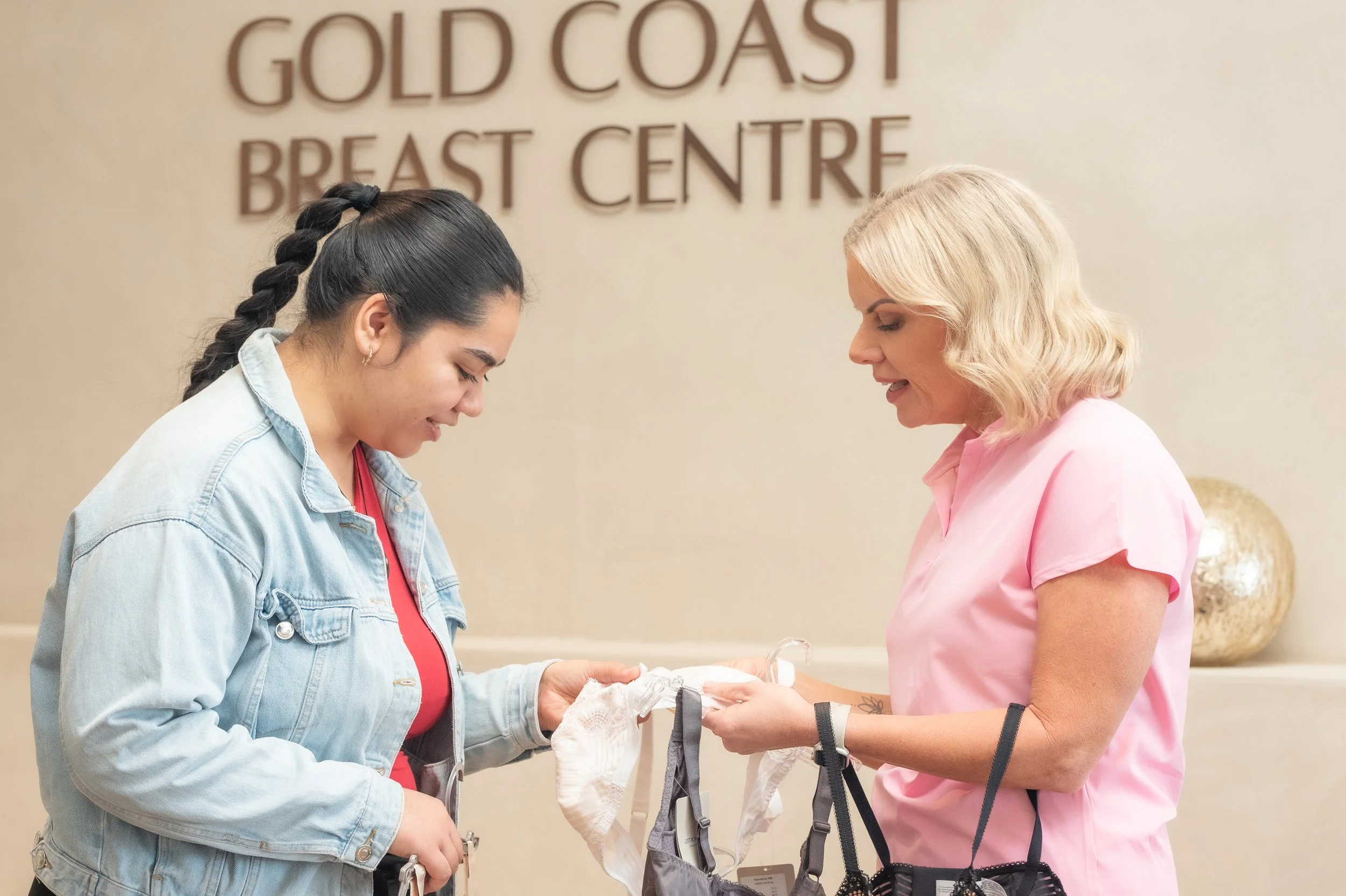 Two women, one with dark hair in a braid wearing a denim jacket and red top, and the other with blonde hair in a pink shirt, are looking at baby clothes in a store with a sign that says "GOLD COAST BREAST CENTRE" in the background.