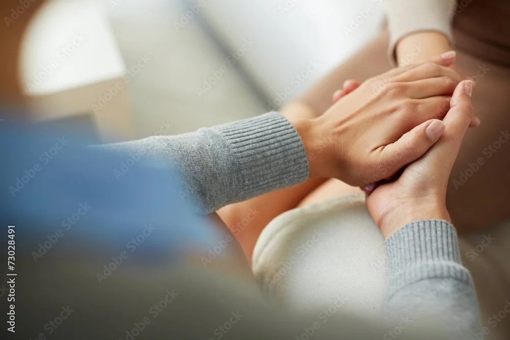 Close-up of two people holding hands, one with a gray sweater and the other with a beige garment, in a comforting gesture.