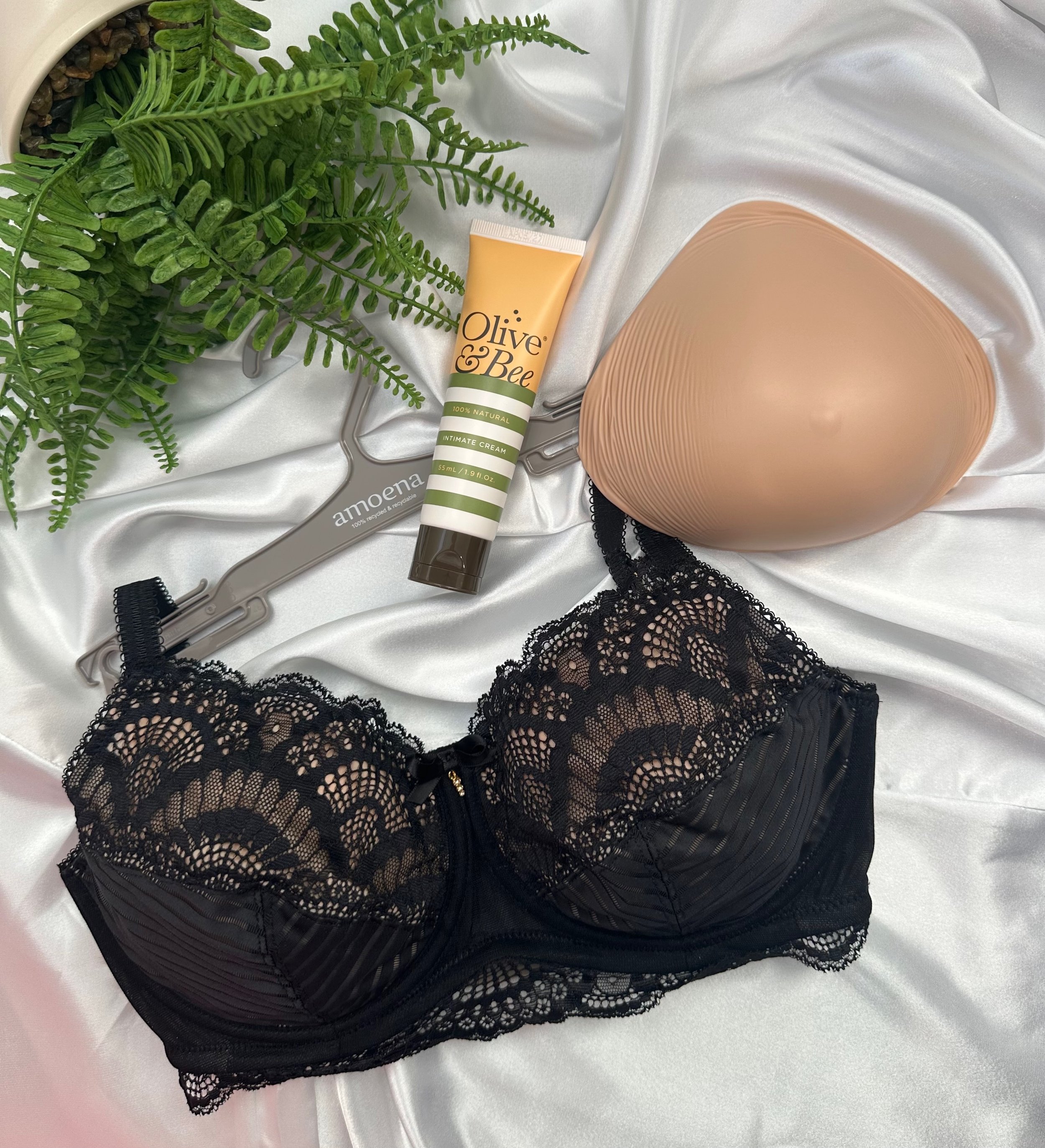A black lace lingerie piece placed on a white satin fabric, with a tube of olive and bee intimate cream, a green fern plant, and a beige silicone device nearby.