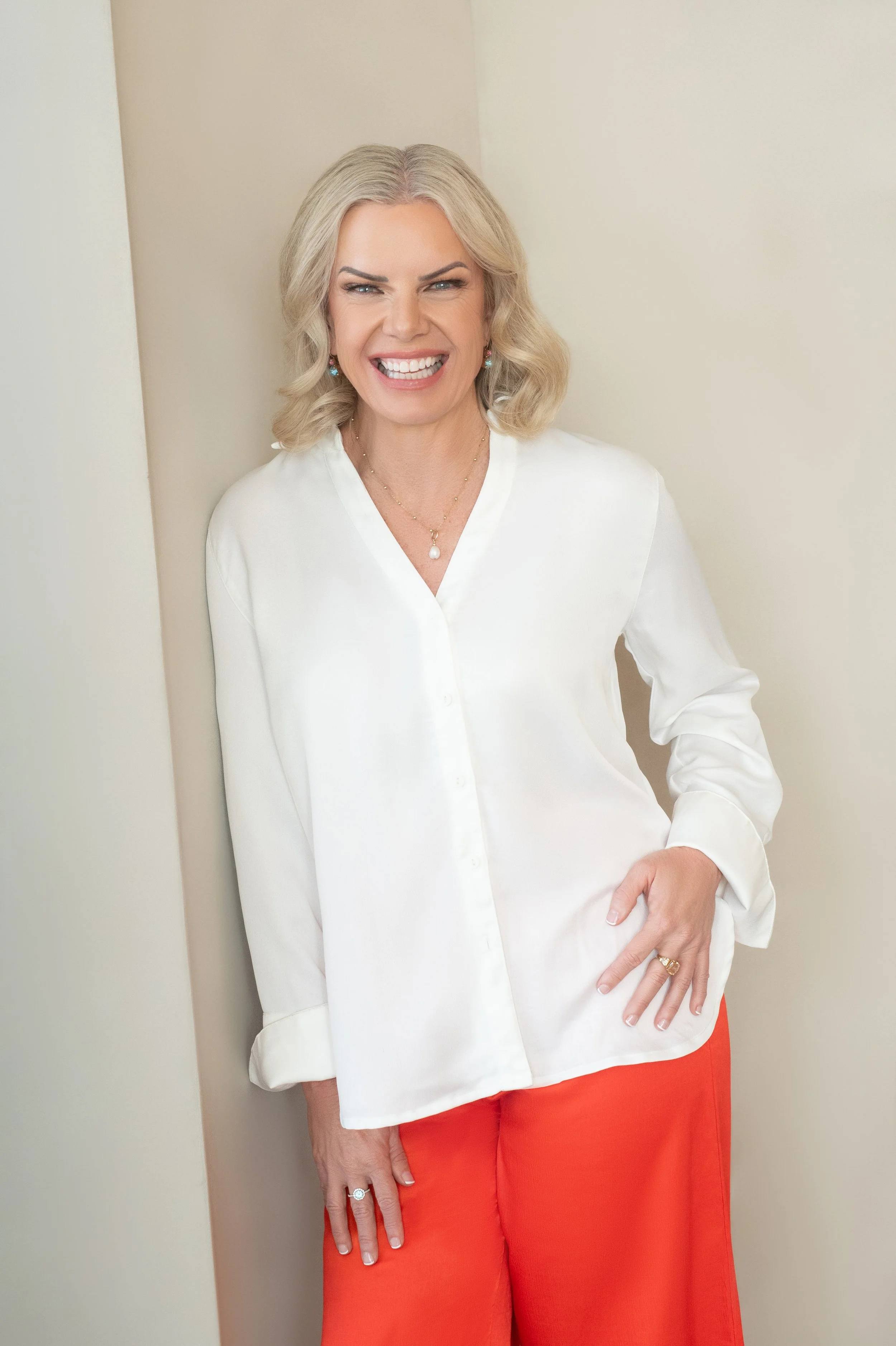 A woman with blonde hair smiling, wearing a white blouse and orange pants, standing against a light-colored wall.