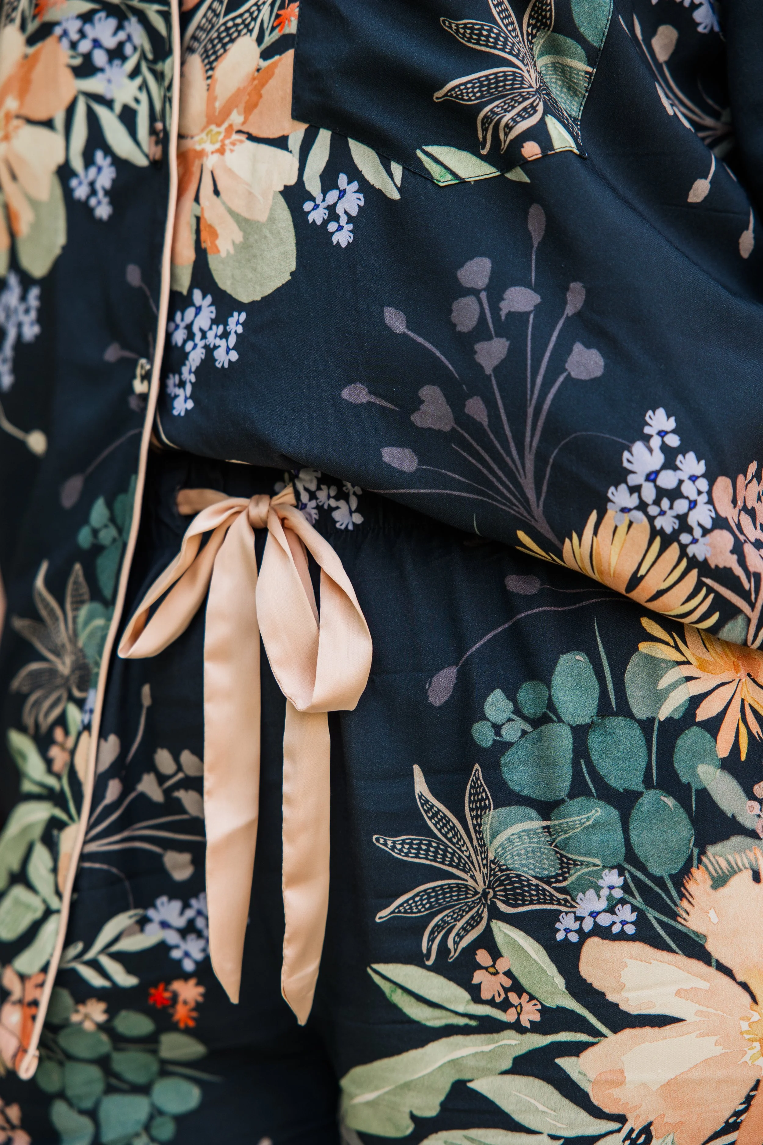 Close-up of a floral-patterned fabric, possibly from a garment, featuring navy blue background with peach, green, white, and pastel-colored flowers and leaves, tied with a light beige ribbon.