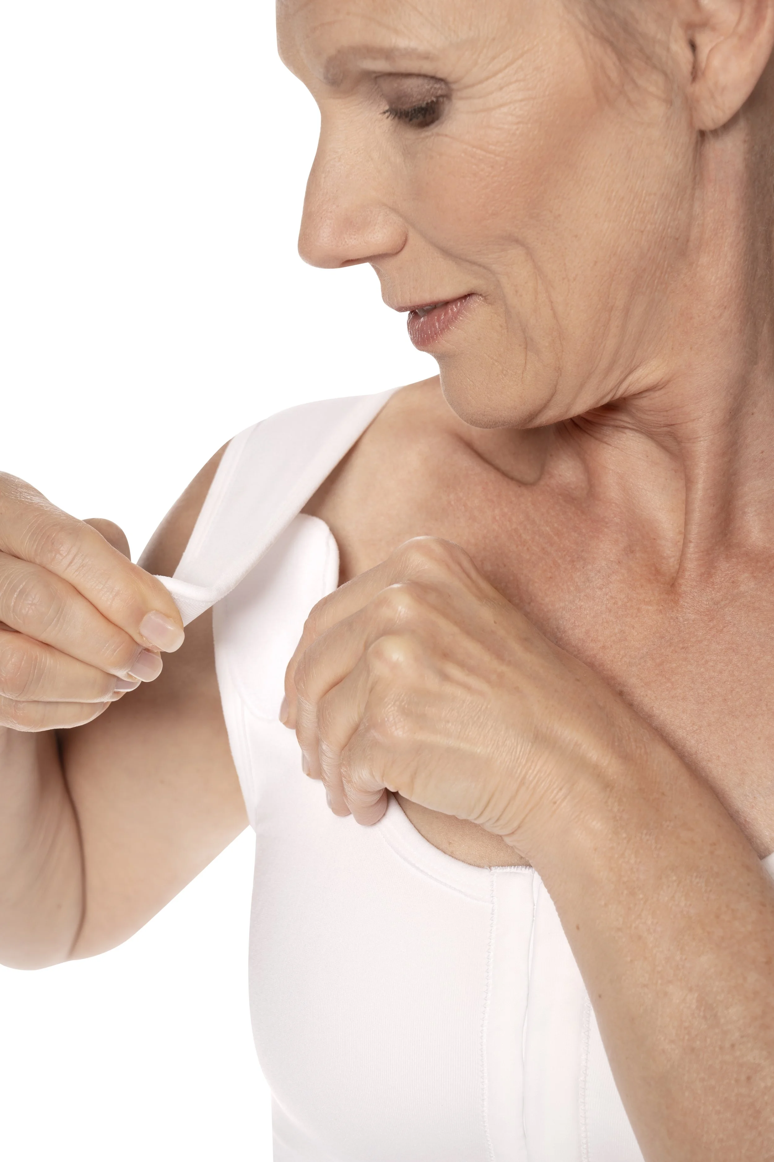 A woman wearing a white tank top is helping herself put on a white bra strap, with a close-up of her upper body and face, showing textured skin and a neutral background.