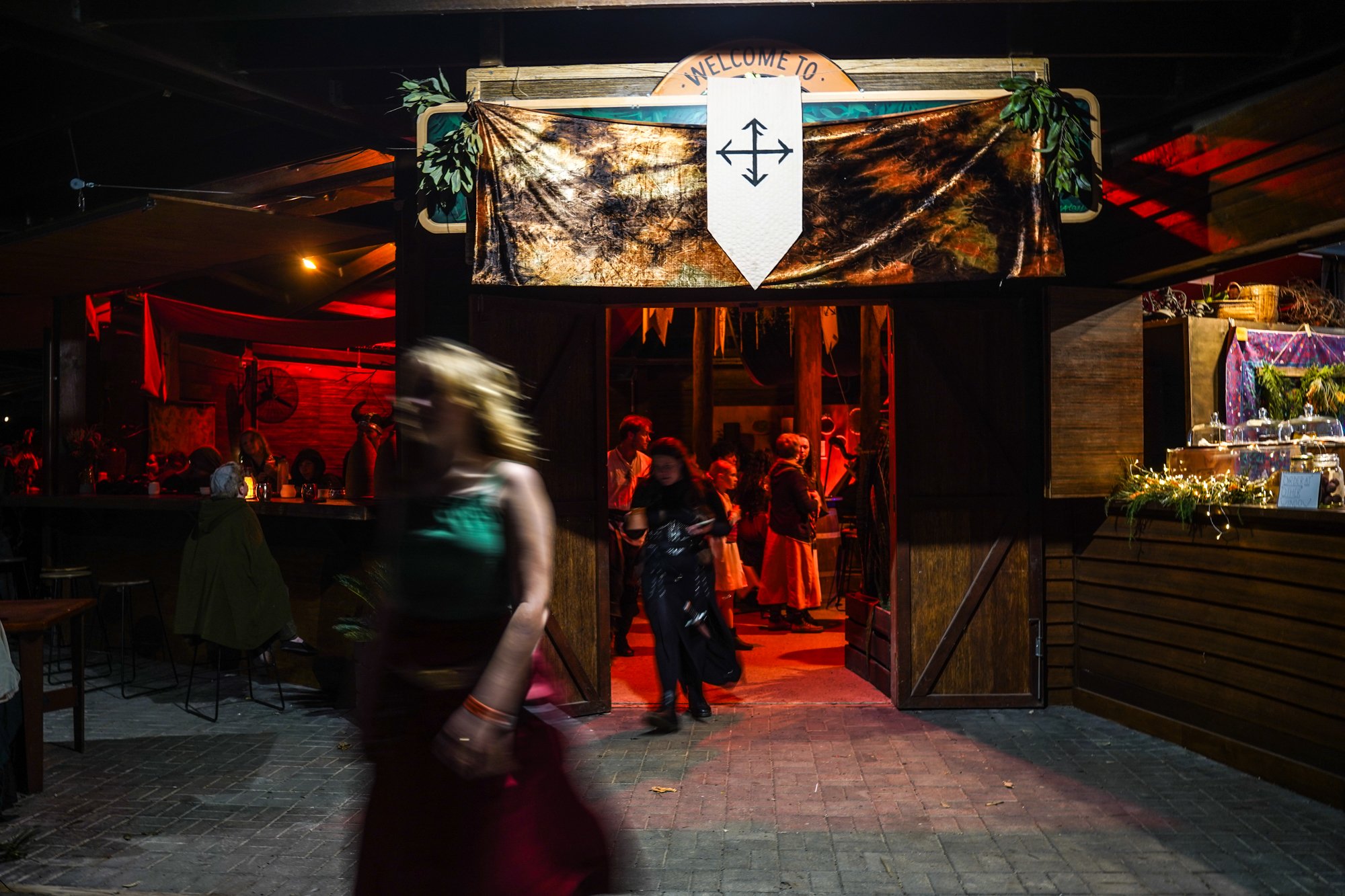 People entering a dimly lit bar or restaurant at night, with a sign that reads 'Welcome to' above the entrance, decorated with greenery and a graphic arrow pointing down.