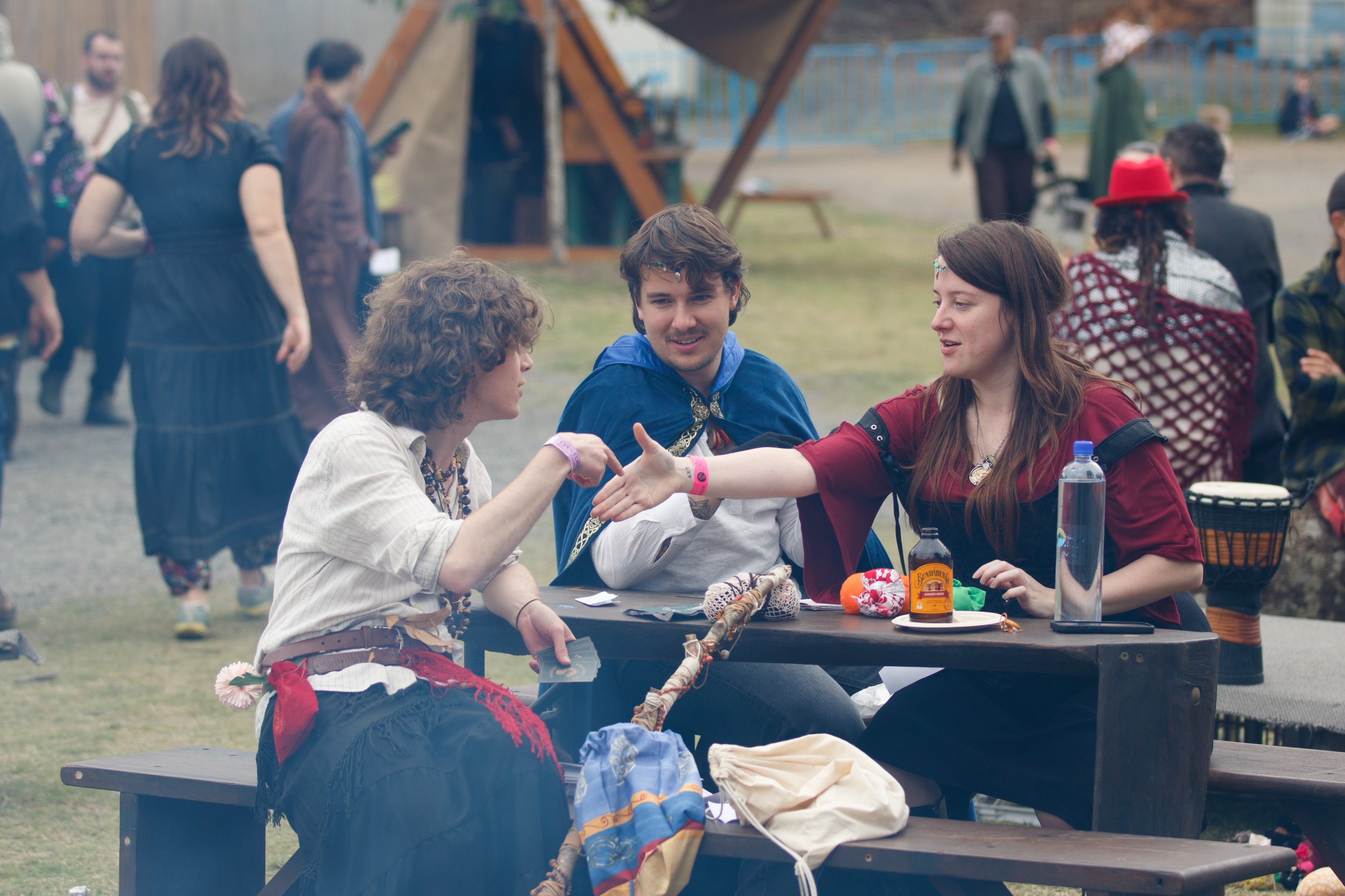 Three people sitting at a picnic table outdoors, engaged in conversation, with other individuals standing in the background at a festival or fair.