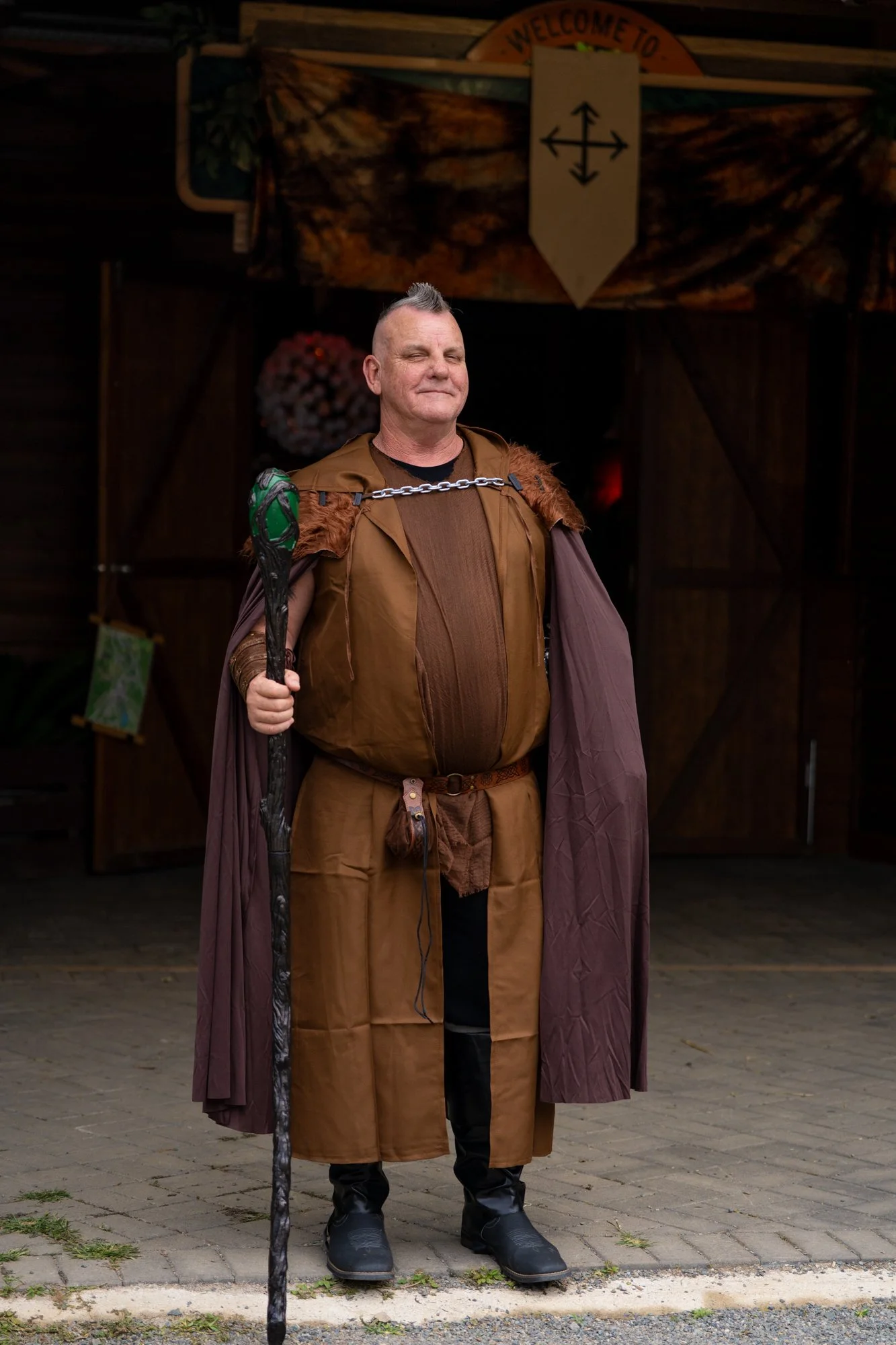 A man dressed as a medieval or fantasy character, wearing a brown and purple cloak, holding a staff, standing outside on a paved area with a wooden building behind him. There is a sign above with a symbol and the words 'Welcome To'.