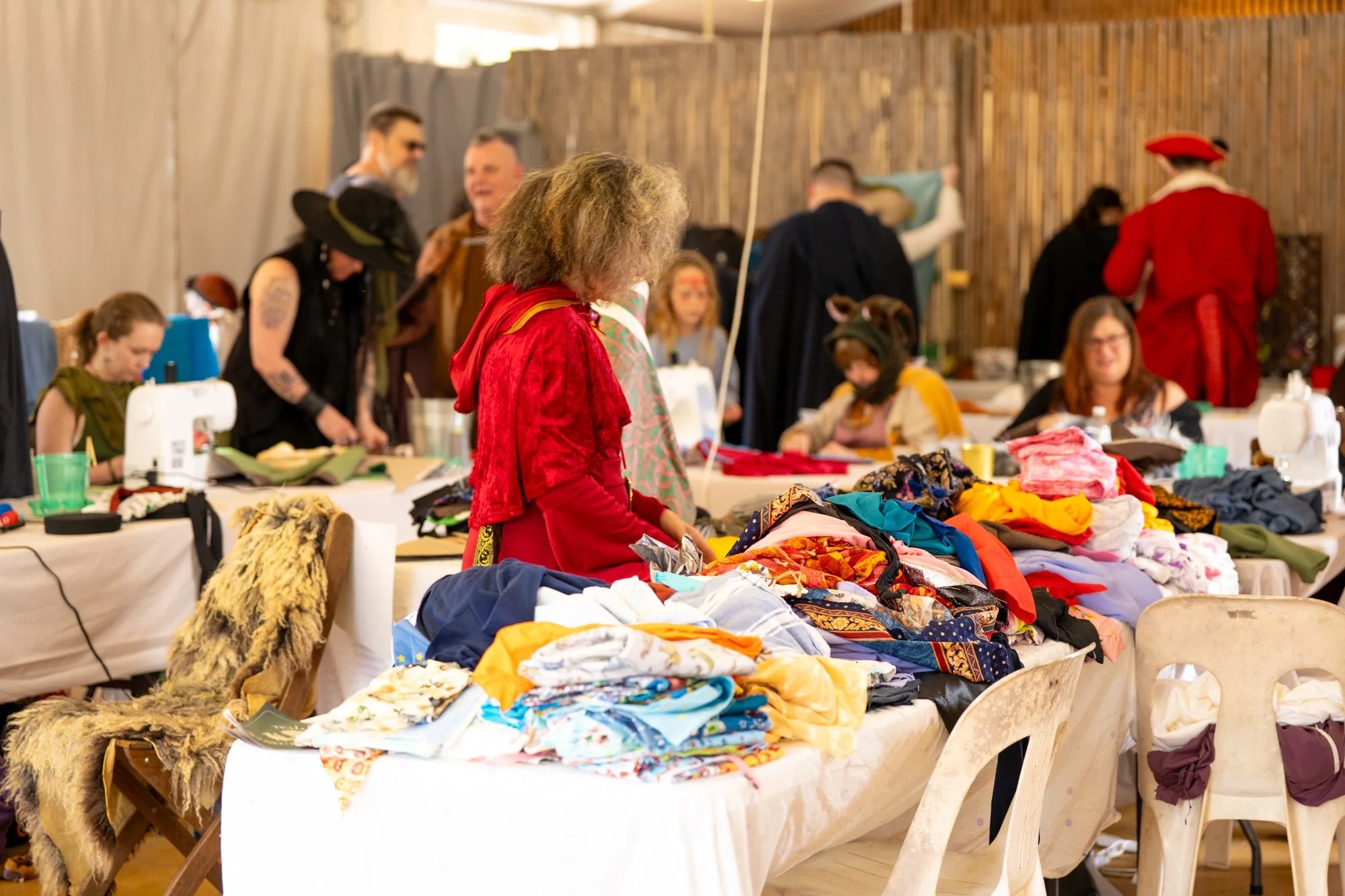 People browsing and shopping at a thrift or flea market with tables filled with clothes, fabrics, and textiles, and a wooden wall in the background.