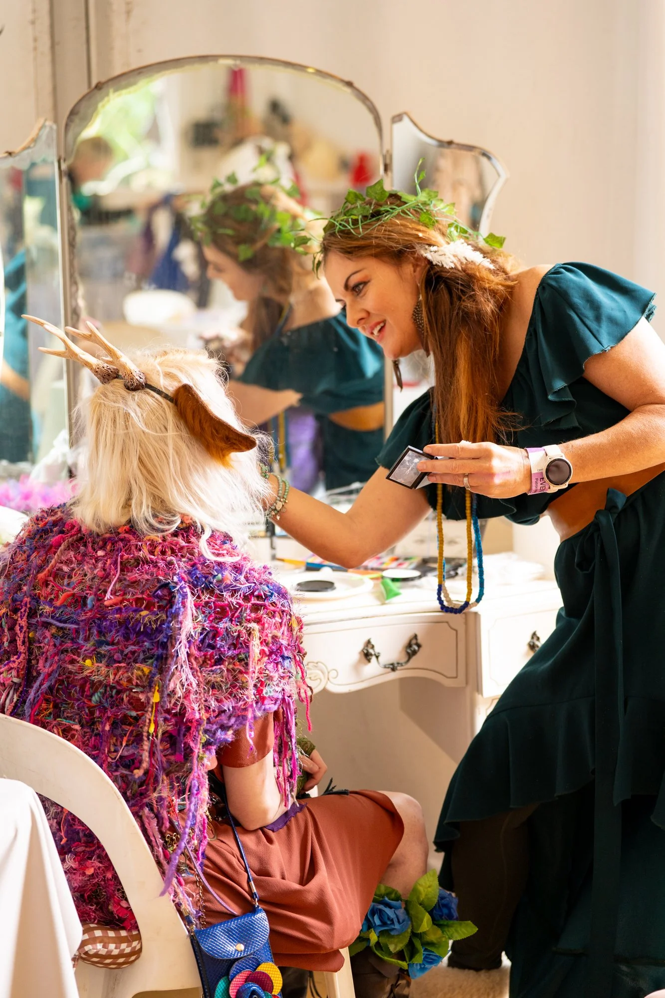 A woman with long red hair dressed as a forest elf is applying makeup to a seated woman with a colorful furry cape and antlers headband, reflected in a mirror behind them.