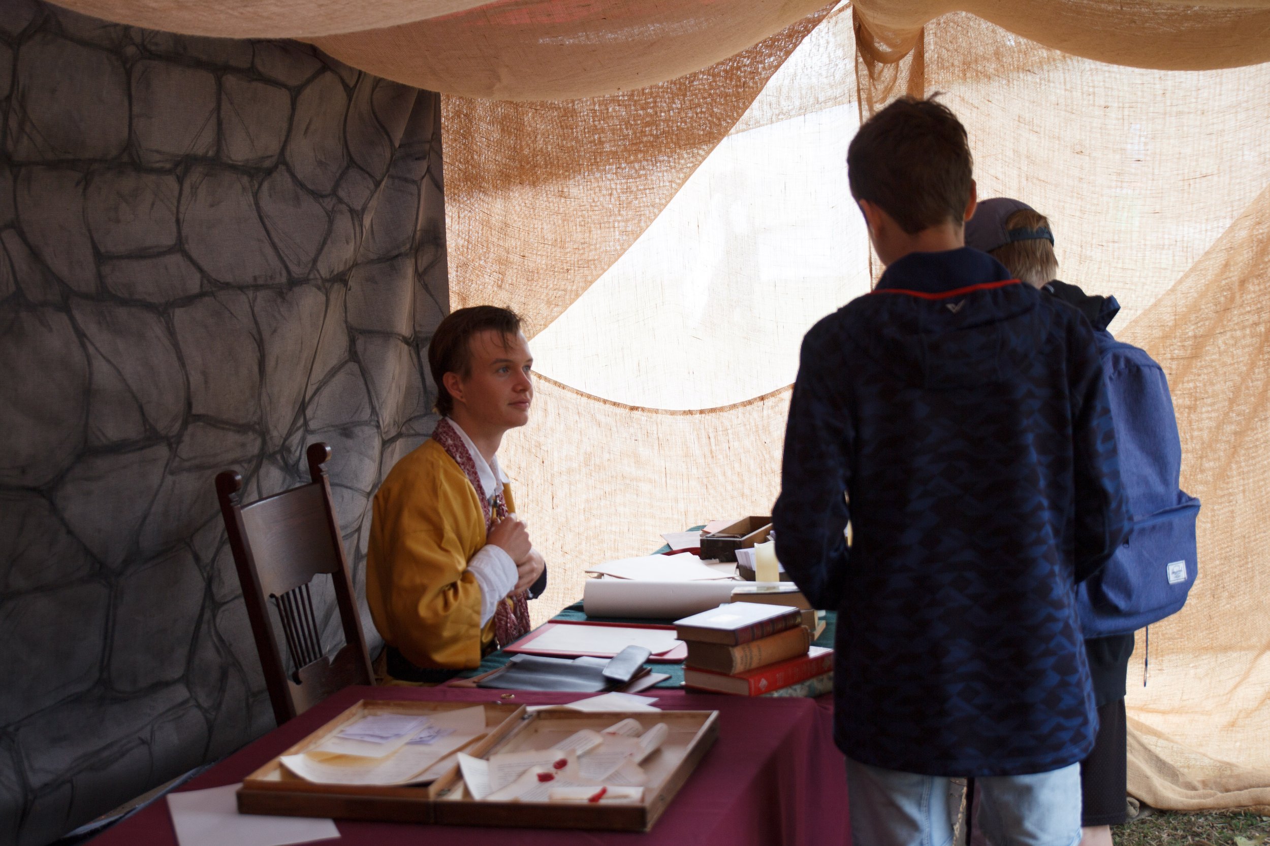 A young woman and several men are engaged in conversation at a table covered with books, papers, and documents, inside a tent with brown fabric walls.