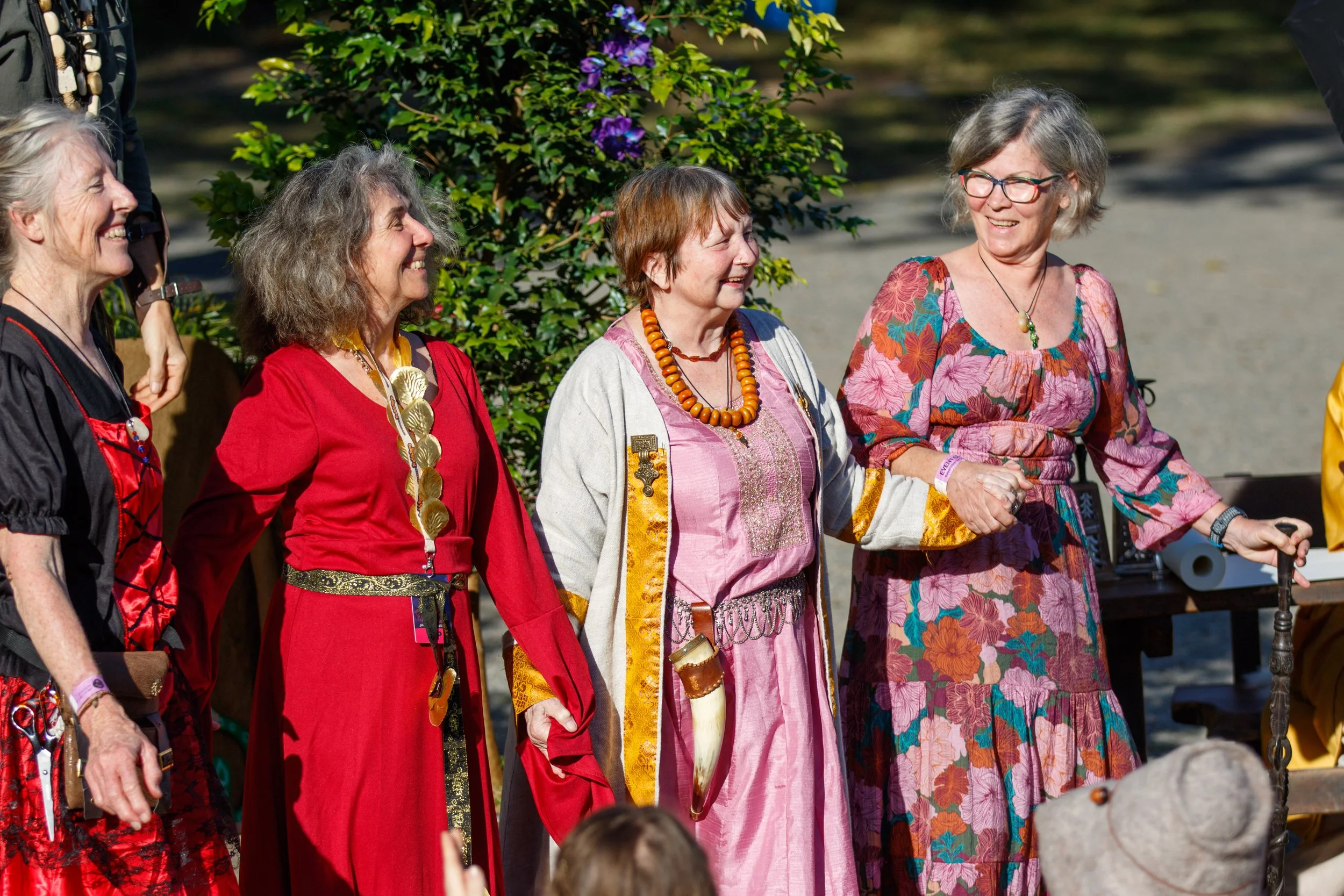 A group of middle-aged women dressed in colorful traditional or folk costumes, standing outdoors in front of green foliage, smiling and appearing to enjoy a social event or celebration.