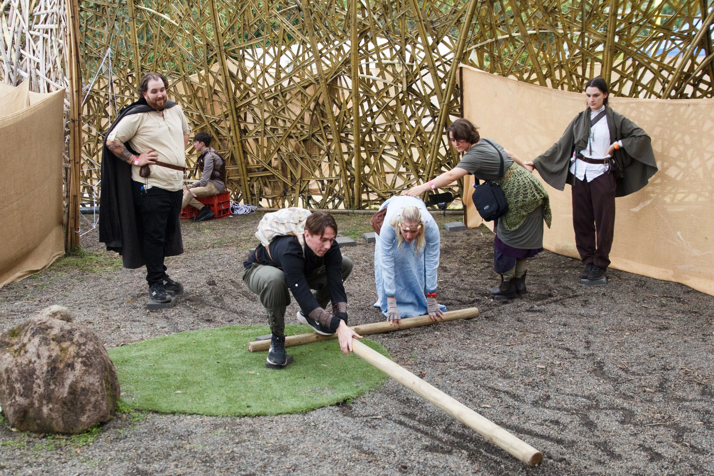 A group of people participating in an outdoor activity involving balancing on log beams in front of a wooden structure made of interwoven bamboo or wood.