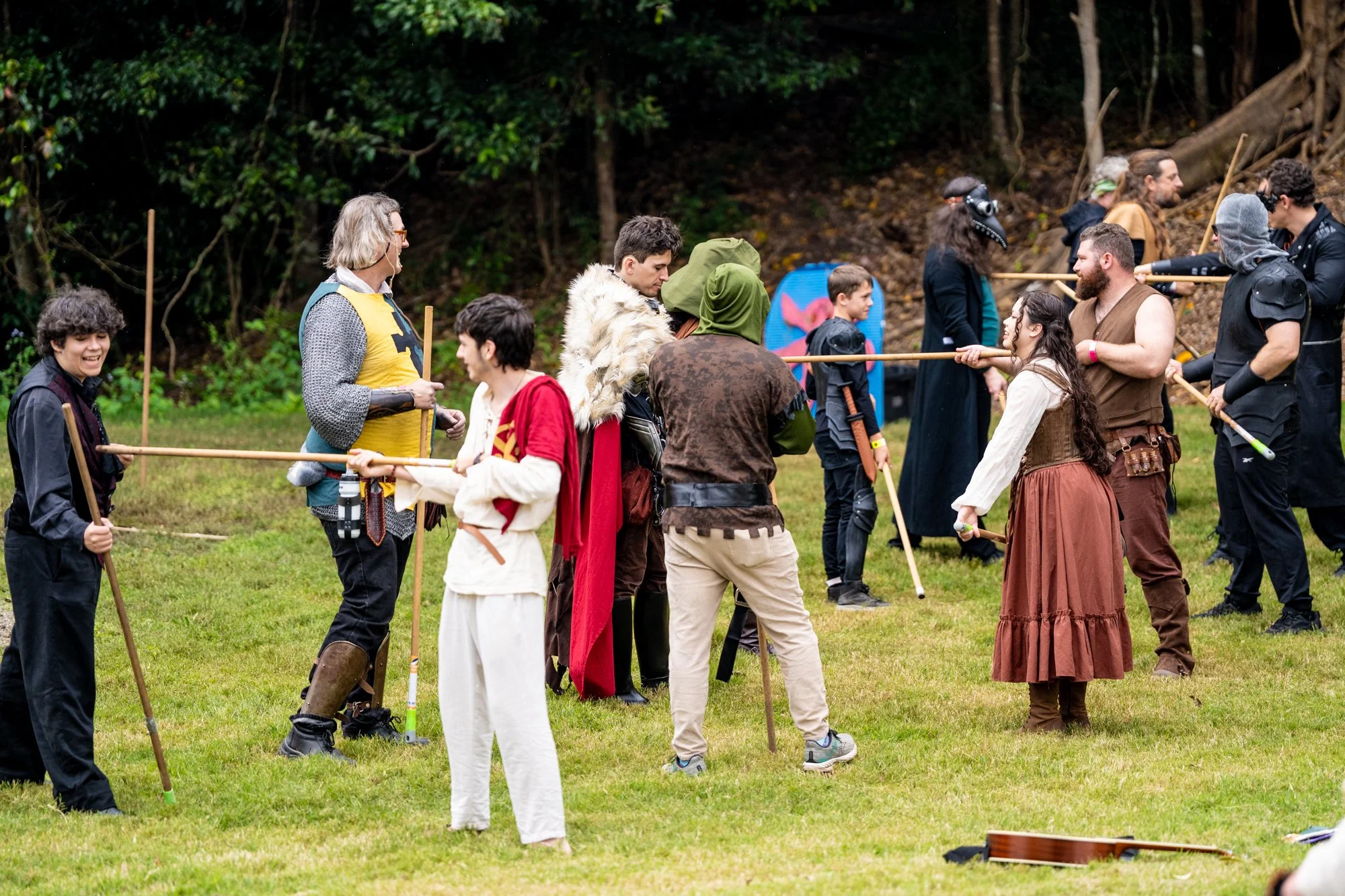 Group of people dressed in medieval costumes participating in a role-playing event outdoors on a grassy area with trees in the background.