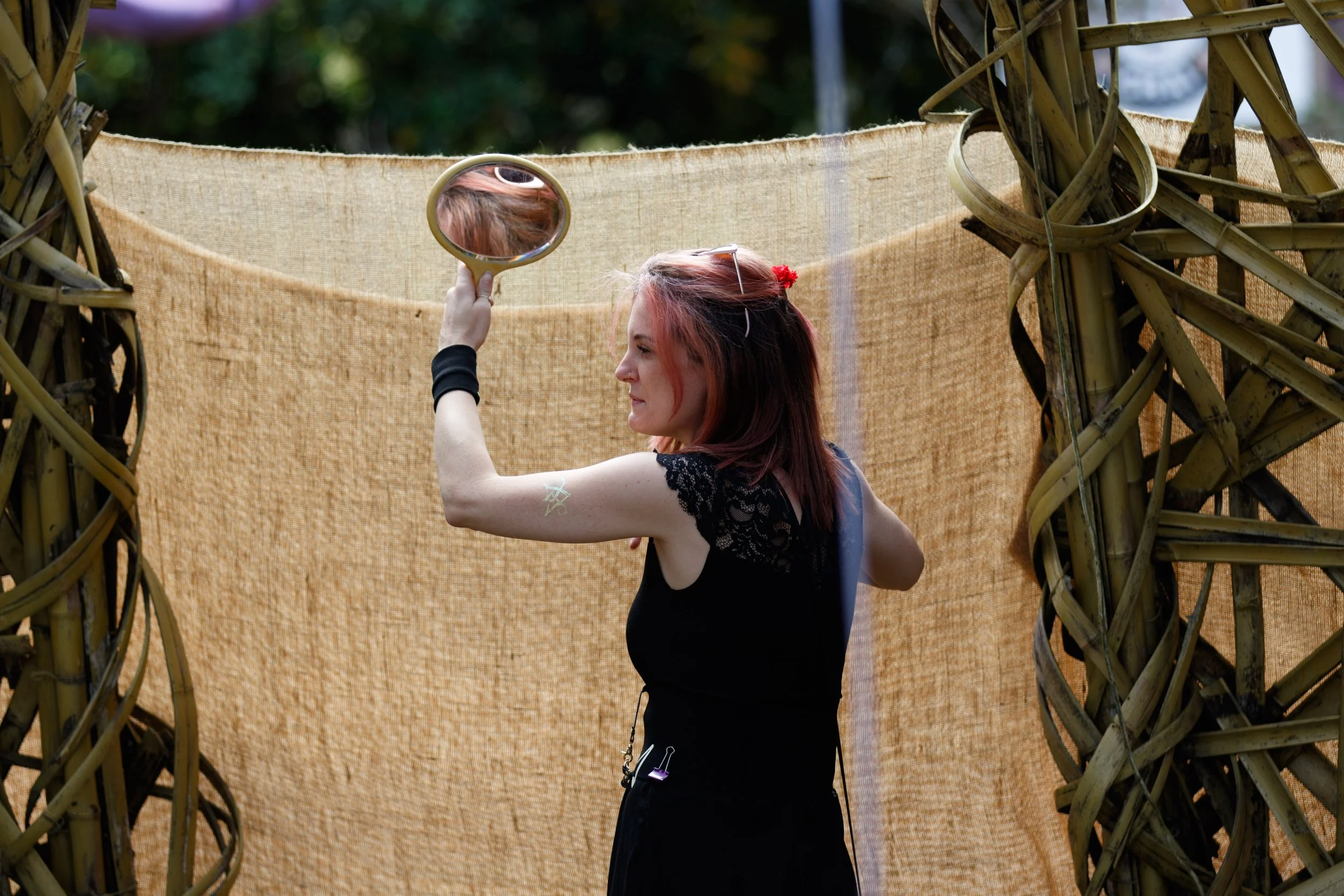 A woman with reddish hair holding a mirror, standing in front of a woven backdrop and a wooden woven arch during an outdoor event.