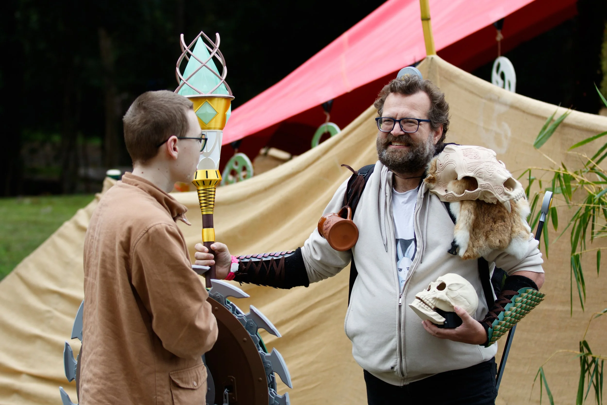 Two men dressed for cosplay or a themed event, standing outdoors near a tent or structure with colorful decorations, engaged in conversation.