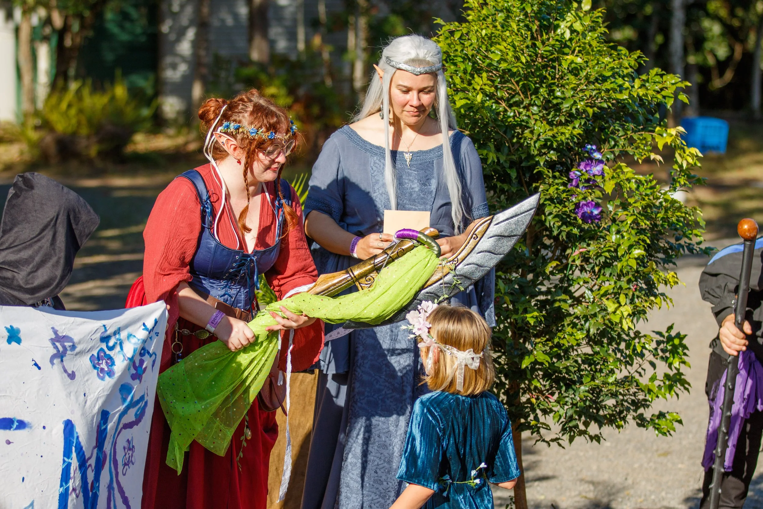 A woman dressed as a fairy or elf with pointed ears and a silver headband, holding a sarong with a child dressed as a fairy in a blue costume, surrounded by other people outdoors during a festival or outdoor event.