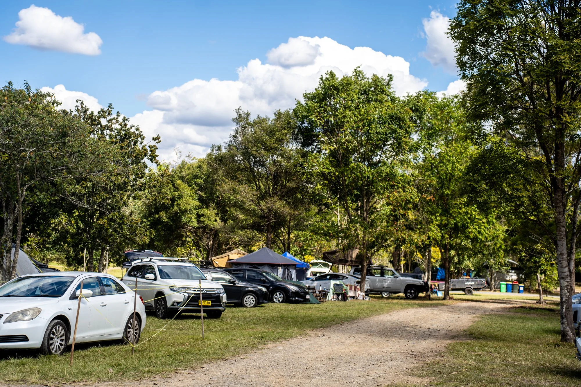 Parking lot with tents, cars, and trees under a cloudy sky.
