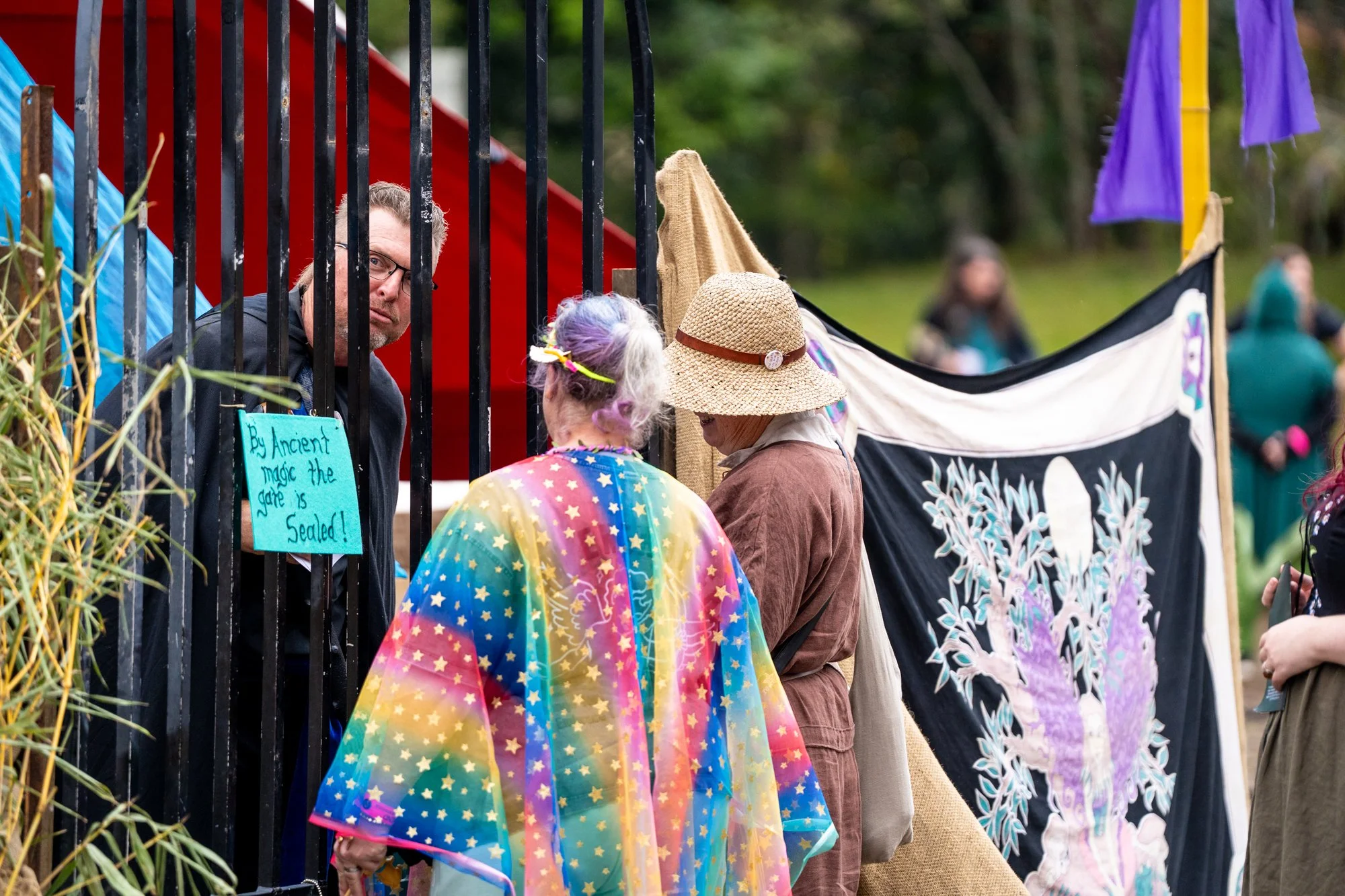 People viewing a person behind a black gate at a fair or outdoor event. One person is wearing a rainbow-colored, star-patterned rain poncho, another has a straw hat, and a third person with purple and white hair is also visible. There are colorful te
