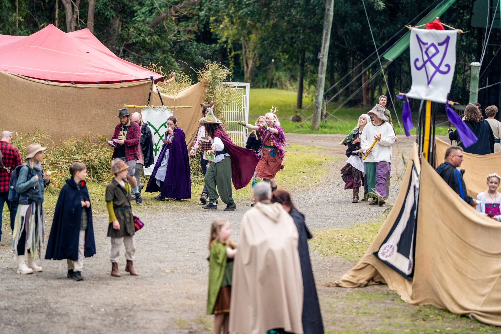 People dressed in costumes, including cloaks, antler headbands, and wizard hats, gathered outdoors near tents with medieval or fantasy festival decor, with trees and power lines in the background.