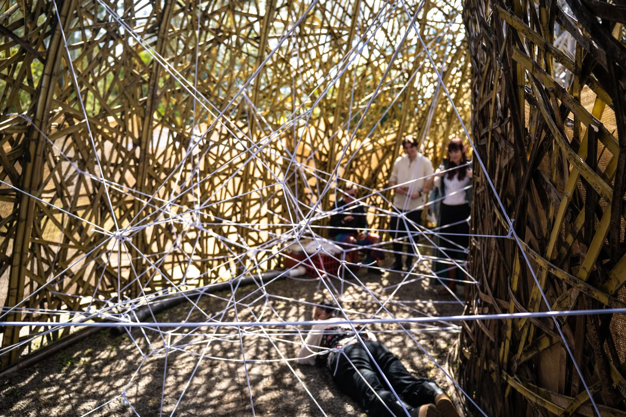 People walking inside a geometric bamboo structure with string lights, with sunlight filtering through.