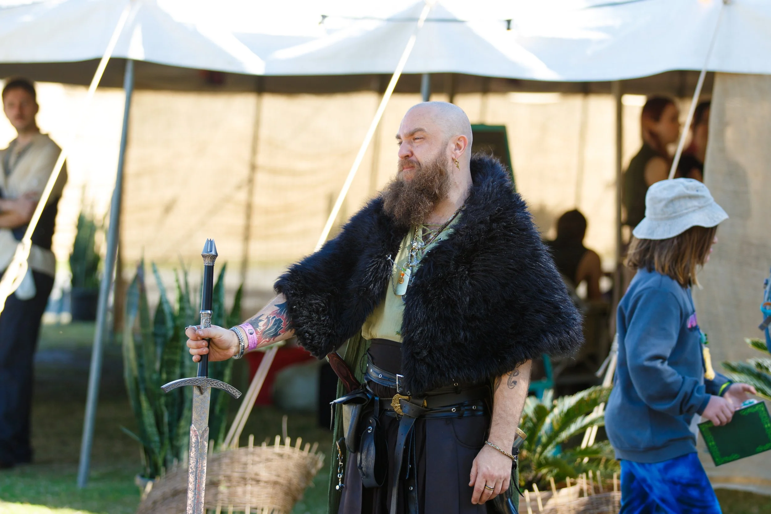 A man with a shaved head and beard wearing a black fur cape and jewelry, holding a sword, standing outdoors at a Renaissance or medieval fair. There are tents and people in the background.
