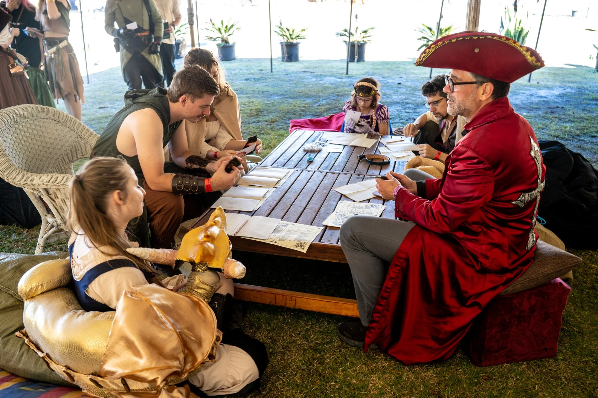 People gathered around a table at a Renaissance-themed event, with some dressed in medieval costumes including a man in a red cloak and a woman in a purple dress with a tiara. The scene appears to be inside a tent or pavilion with outdoor scenery vis