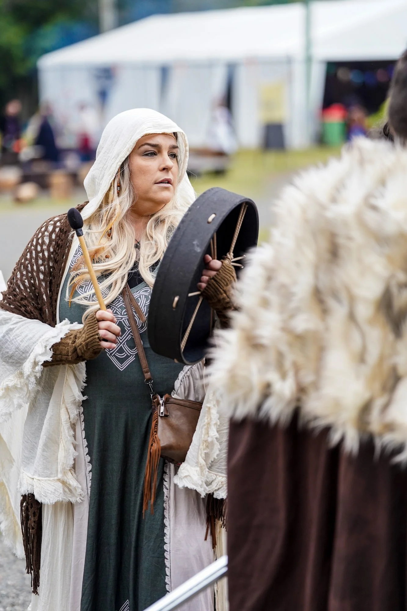 A woman dressed in medieval or fantasy costume, holding a tambourine, standing outdoors at an event with tents and people in the background.