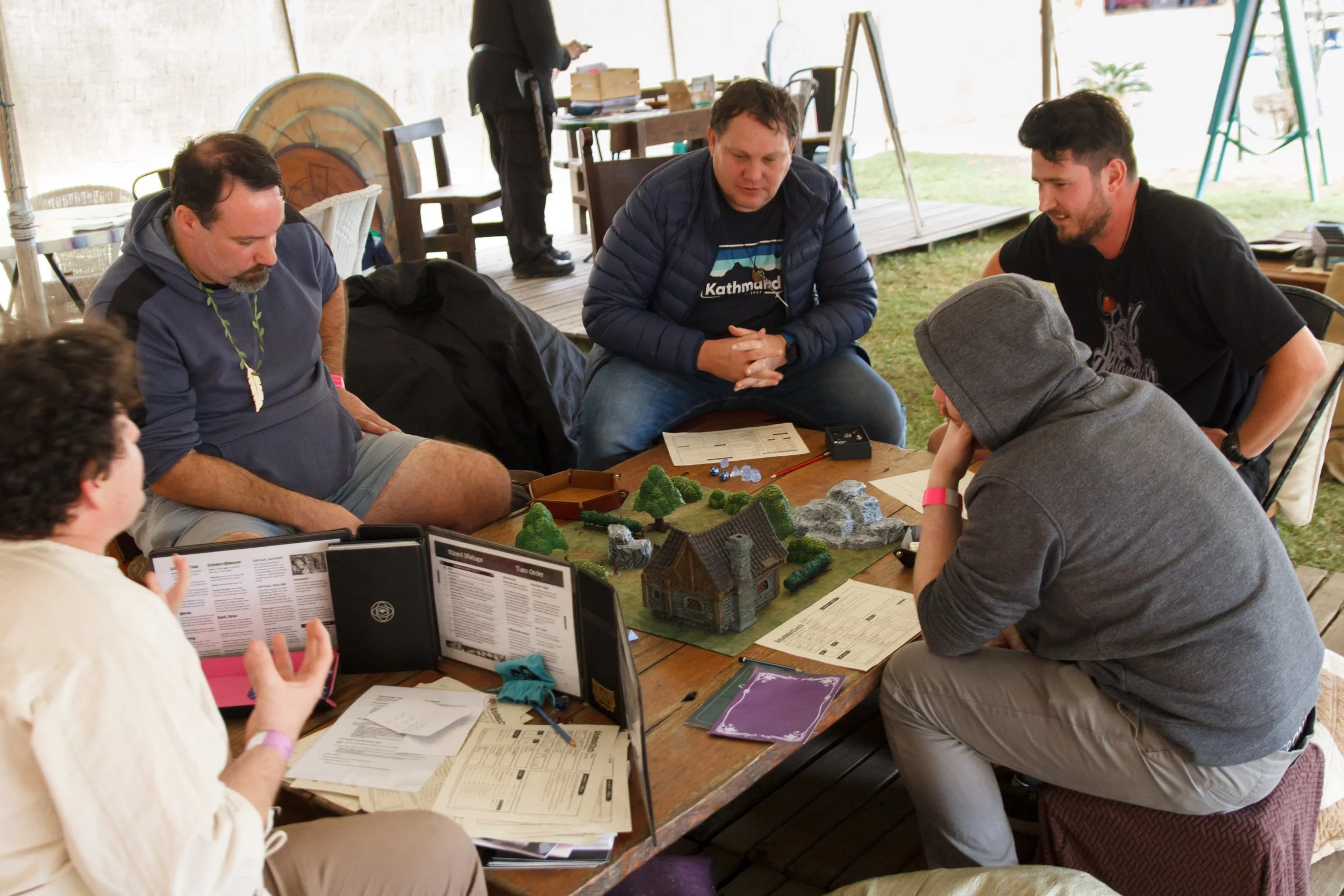 A group of people gathered around a table playing tabletop role-playing game, with character sheets, manuals, and miniature terrain on the table in an outdoor tent.