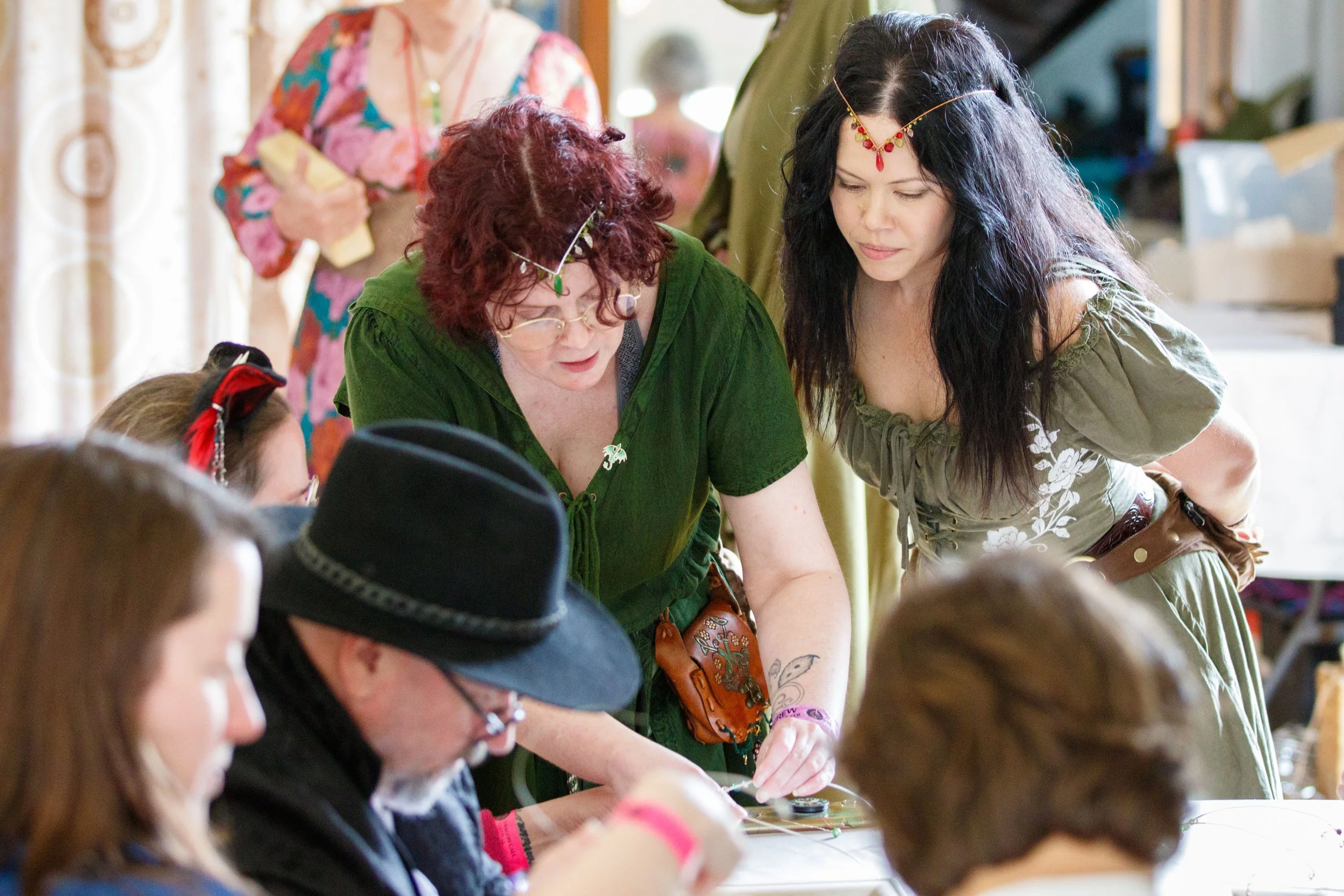 A group of women, some with red and black hair, wearing traditional and modern clothing, are gathered around a table engaging in a craft or art activity inside a room with natural lighting.