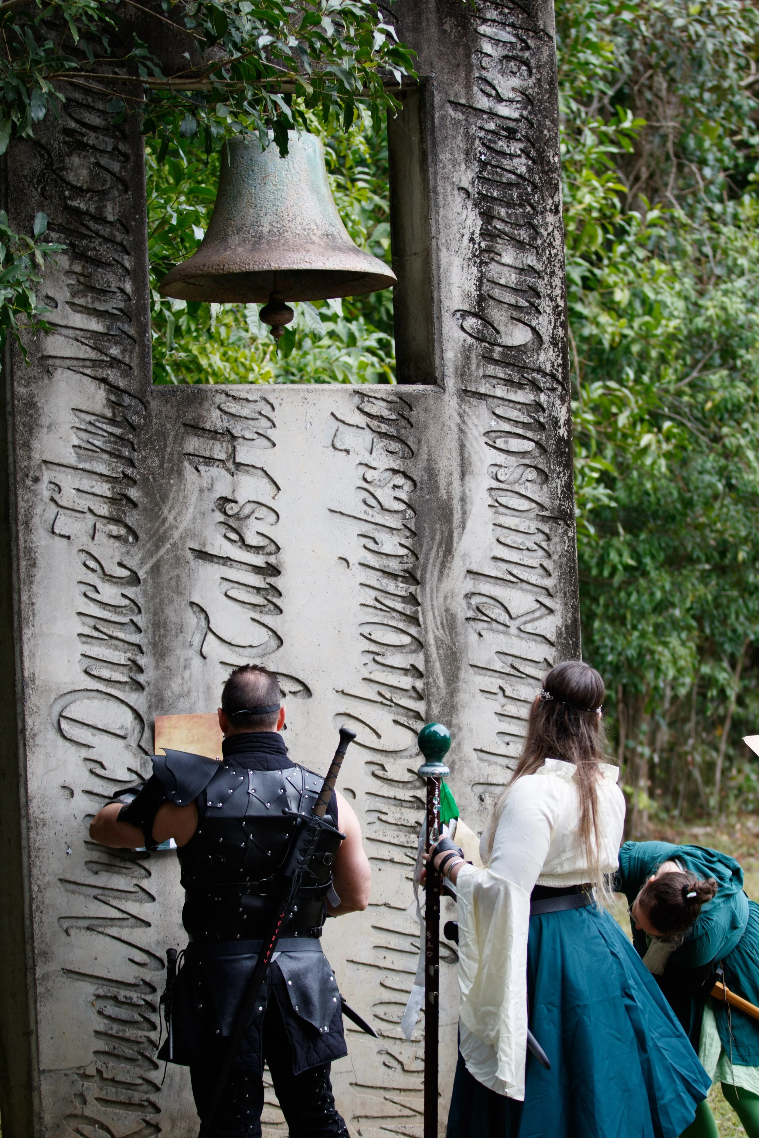 People dressed in medieval costumes gathered around a large stone monument with an engraved inscription and a hanging bell in an outdoor setting surrounded by greenery.