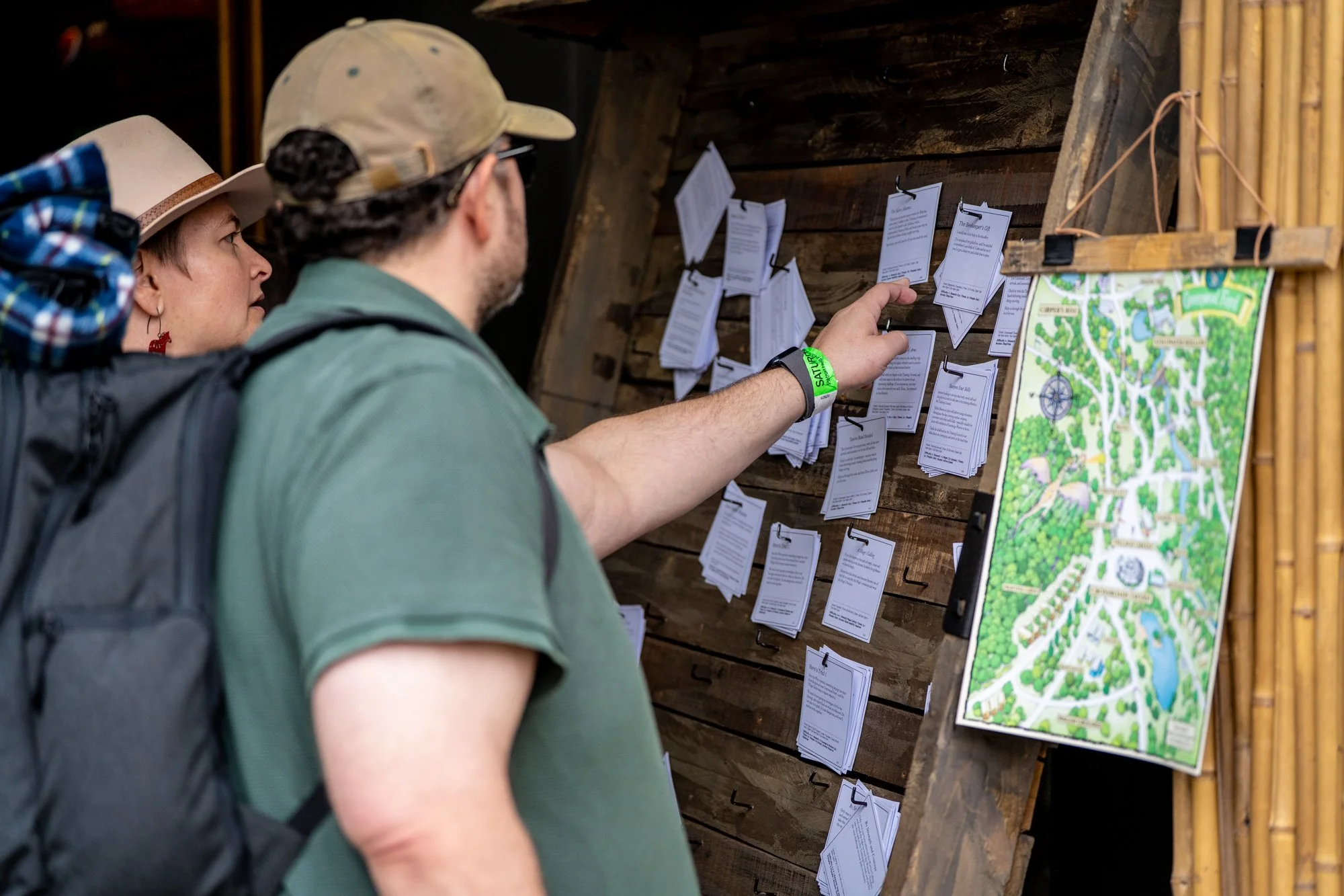 Two people, a woman and a man, are looking at notices and maps posted on a wooden wall in an indoor setting. The woman is wearing a hat and a backpack, and the man is wearing a cap and glasses.