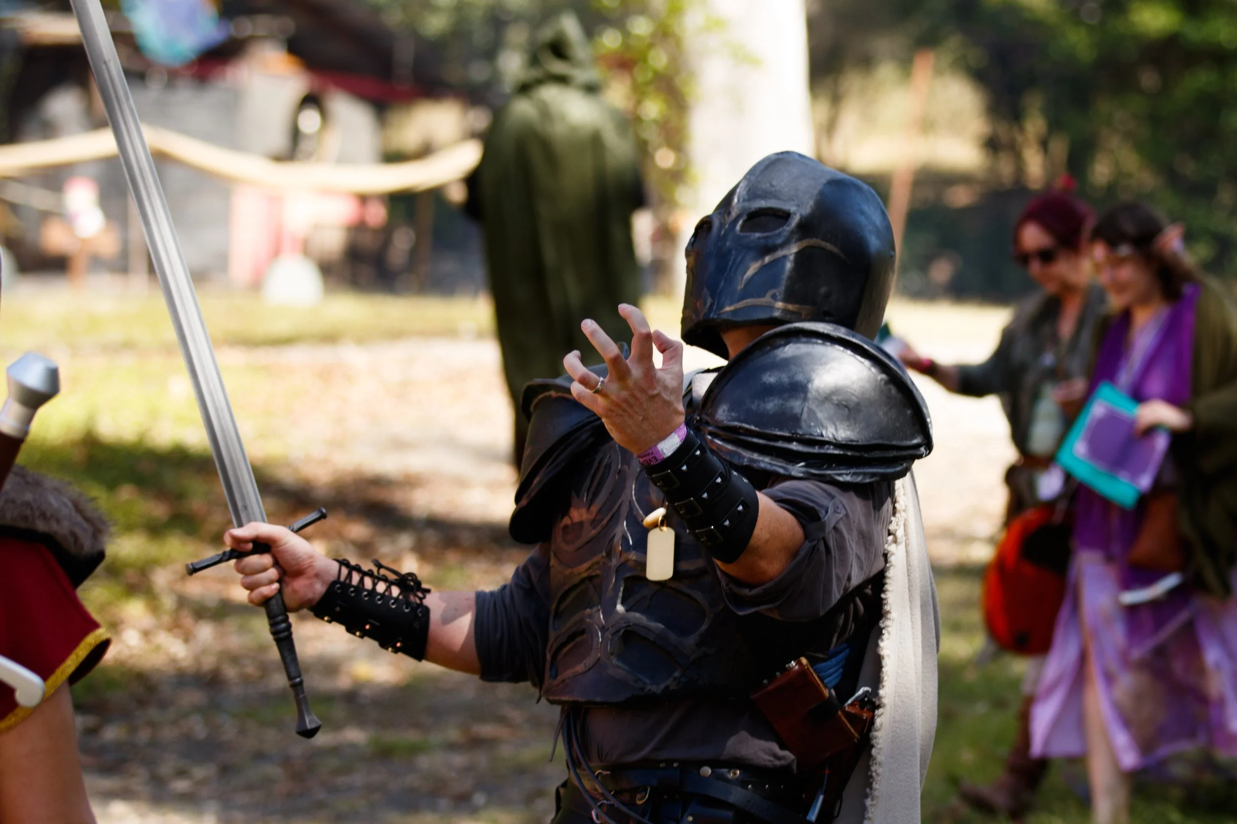 Person dressed in a black knight costume with a helmet and armor-like details, holding a sword, at an outdoor event with several other people in the background.