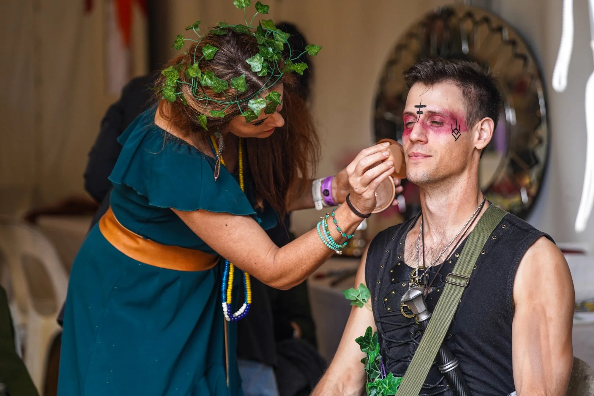 A woman with curly hair wearing a crown of green ivy leaves applies makeup to a young man with face paint, jewelry, and a harness, inside a room with a mirror in the background.