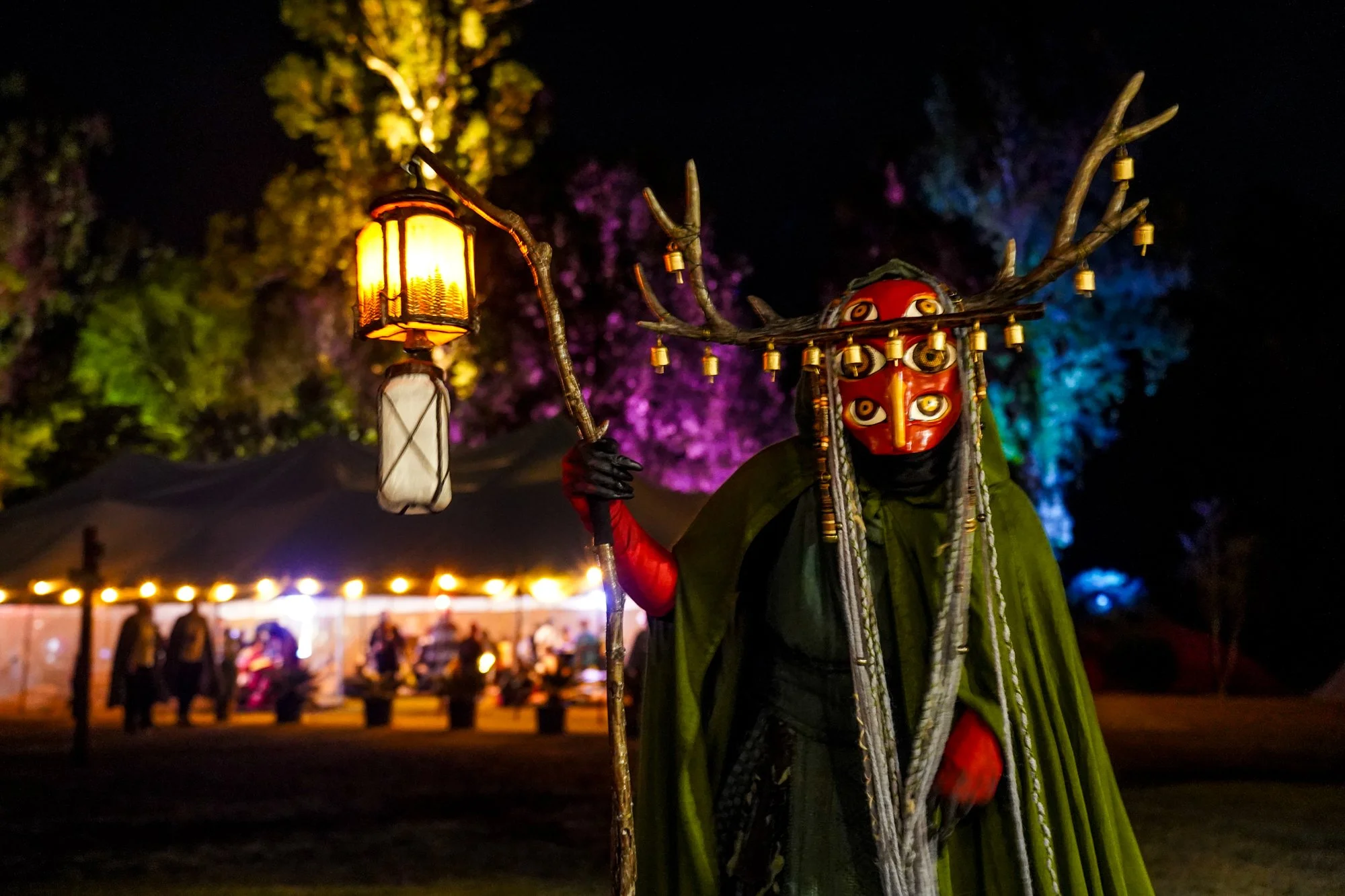 Person dressed in traditional costume with a red mask, wearing a mask with multiple faces, holding a staff with a lantern, and wearing antler-like branches with small lanterns attached, standing outdoors at night with colorful illuminated trees in th