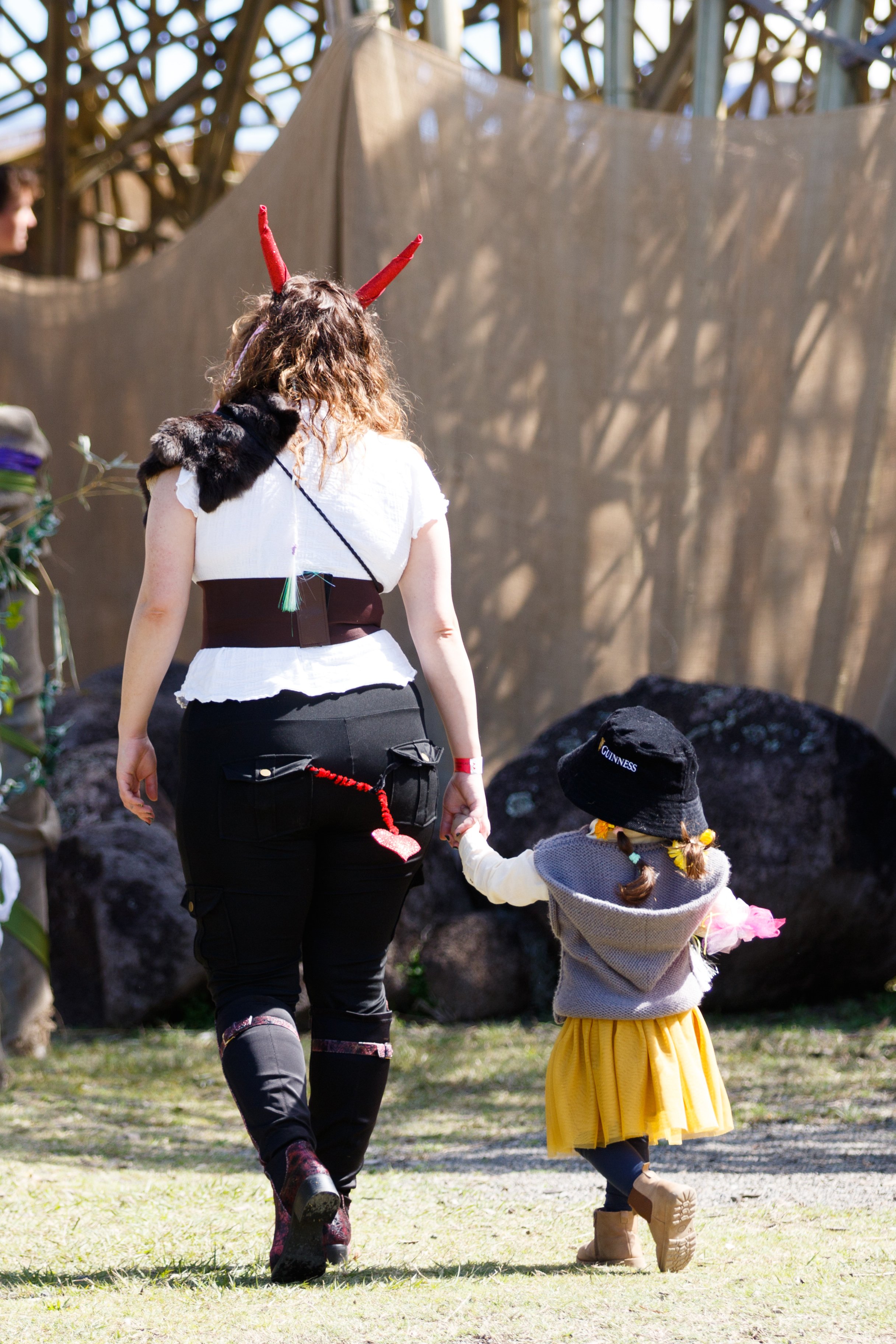 A woman and a girl holding hands and walking outdoors on grass. The woman is wearing a costume with devil horns and a fur shoulder accessory, and the girl is dressed in a yellow skirt, gray hoodie, and boots. There is wooden structure or sculpture in