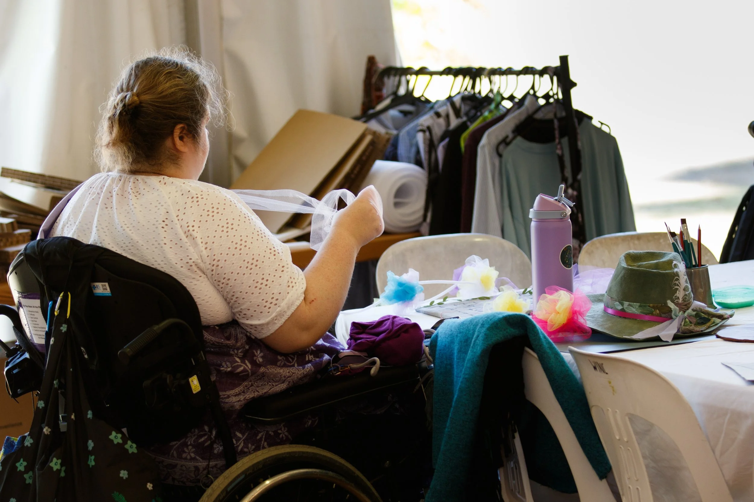 A woman in a wheelchair sitting at a table covered with craft supplies, including colorful tissue paper, cotton, and a pink water bottle, while working on a craft project.