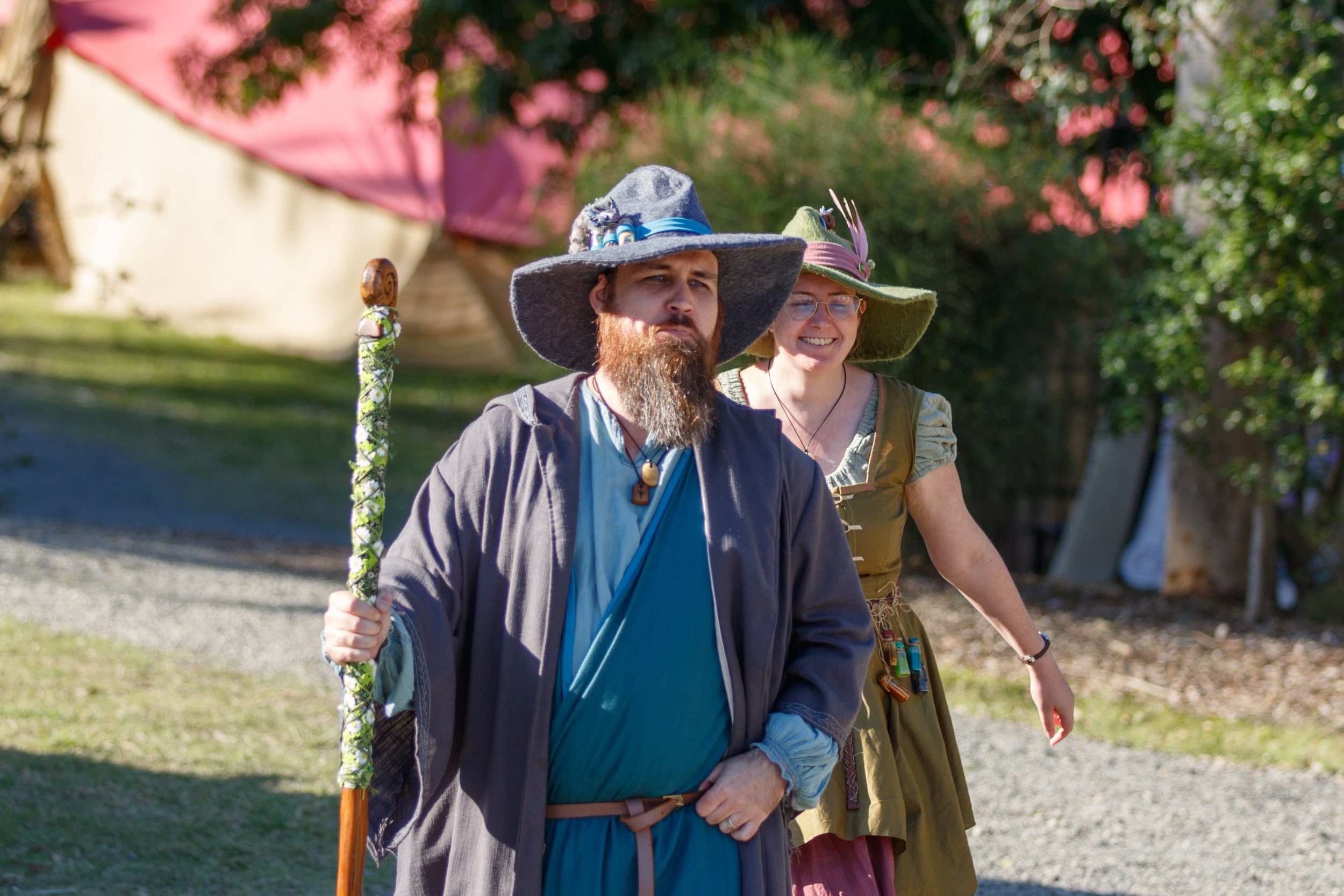Two people dressed as wizards or witches, wearing large pointed hats, walking outdoors in a sunny park or garden, with trees and a pink tent in the background.