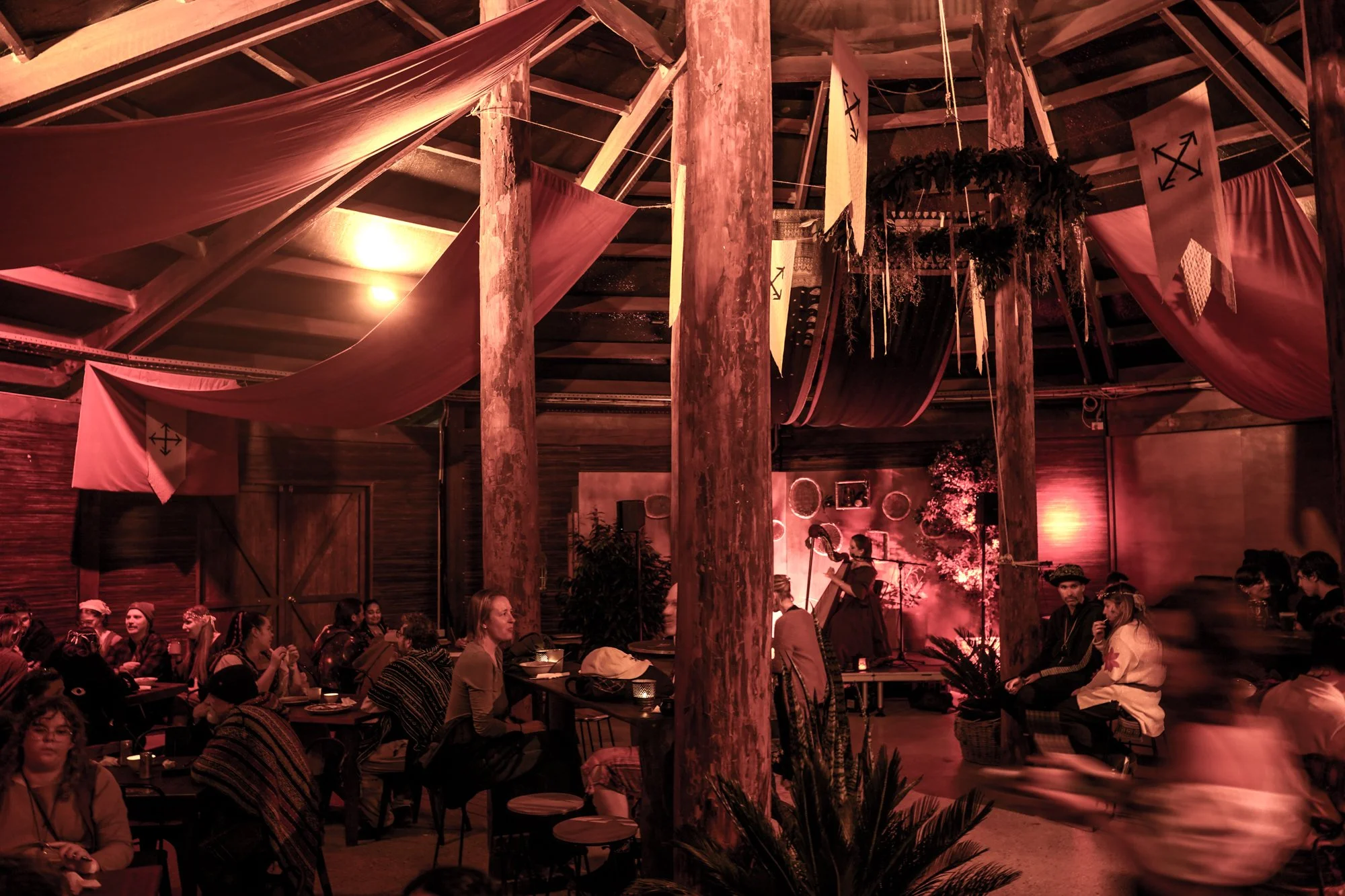 Indoor event with people sitting at tables, wooden beams and curtains decorated with black arrow symbols, warm lighting, and some plants and hanging decorations.