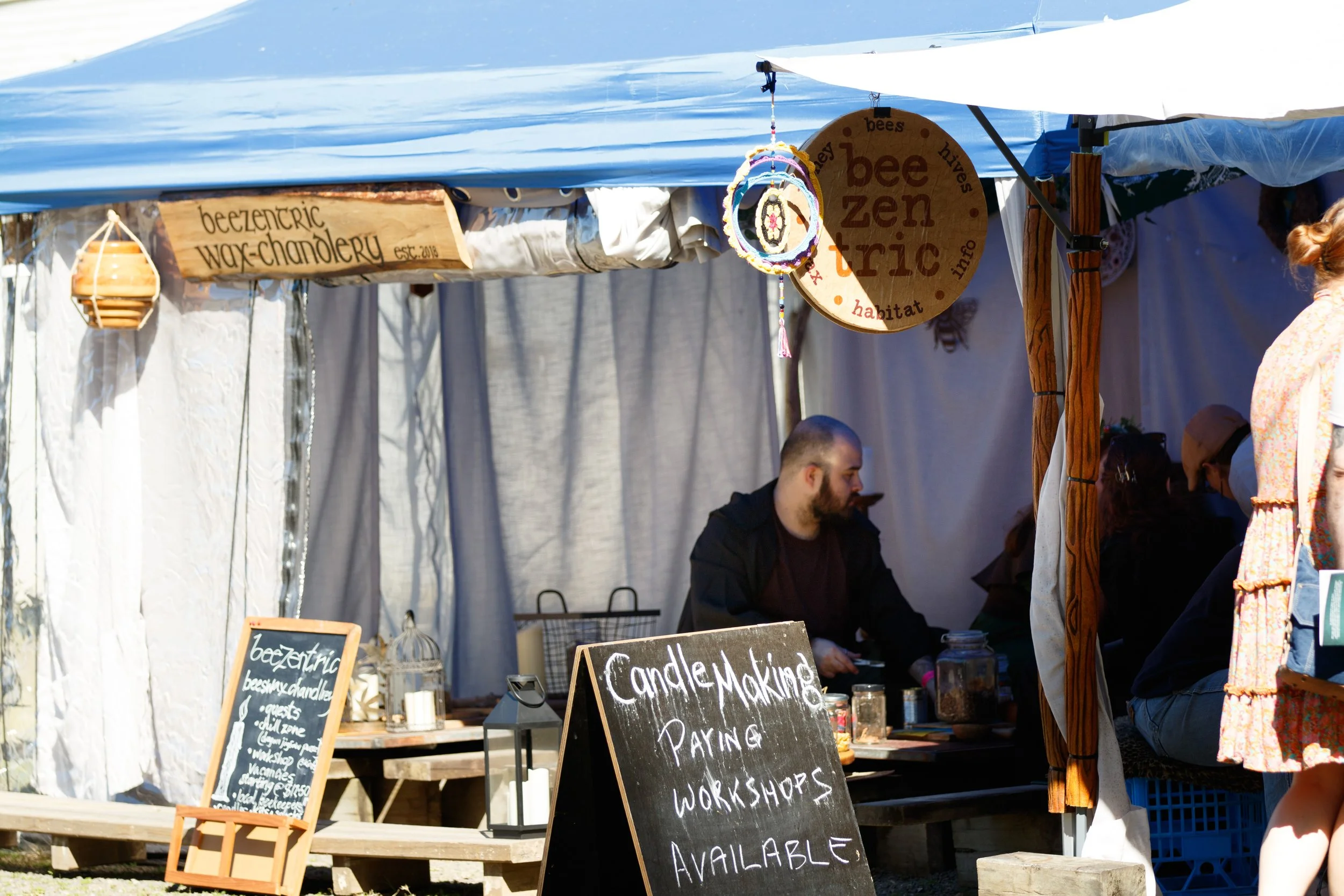 Outdoor market stall with signs advertising beeswax candles, candle making, and workshops. Several people are visible browsing or working behind the counter, with a white canopy overhead.