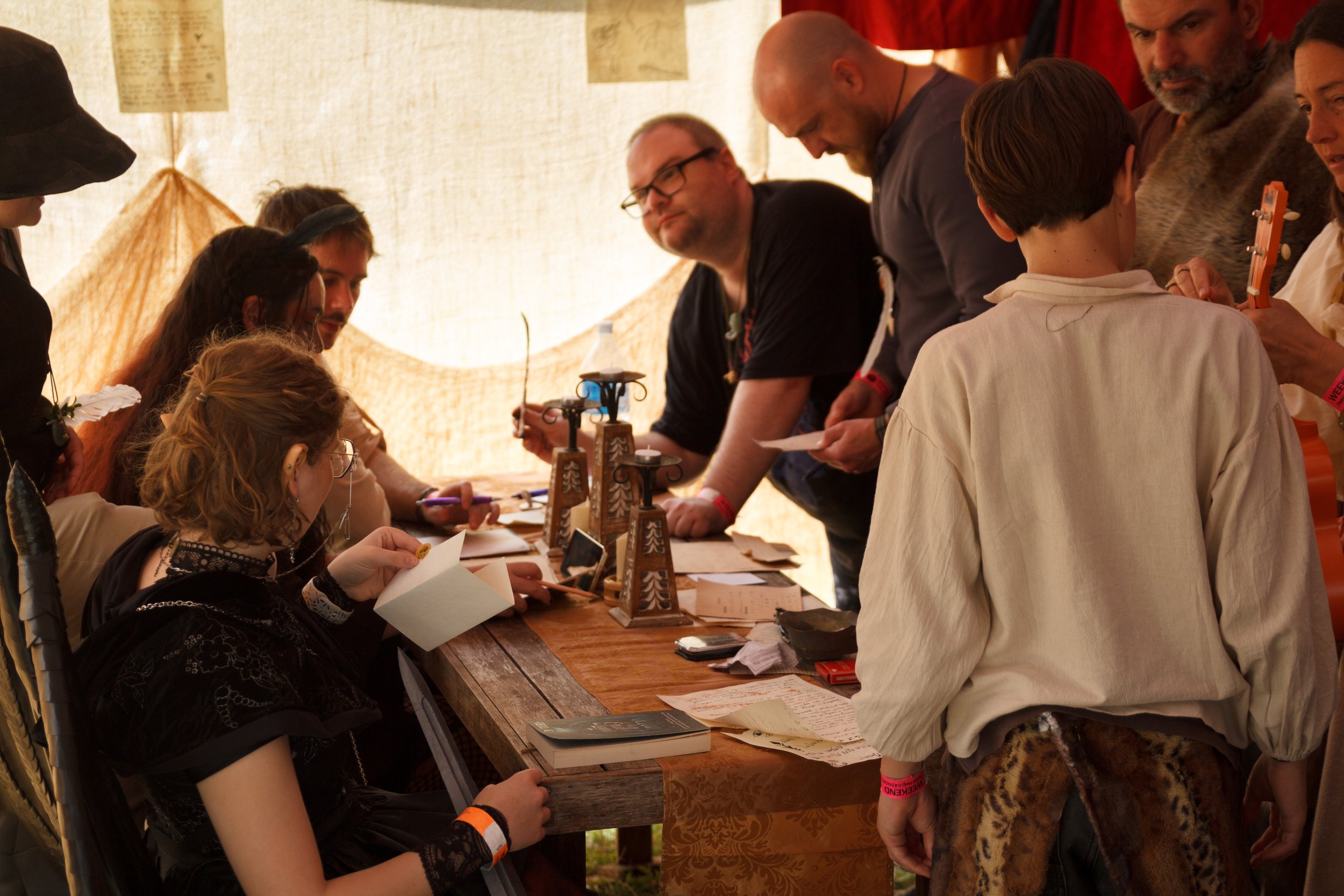Group of people dressed in medieval or fantasy costumes gathered around a wooden table, engaging in a game or activity inside a tent or pavilion.
