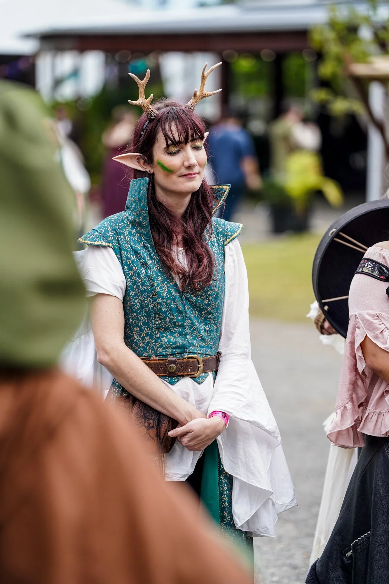 A woman dressed as a fantasy elf with antlers, pointed ears, and face paint, standing with her eyes closed during an outdoor event.