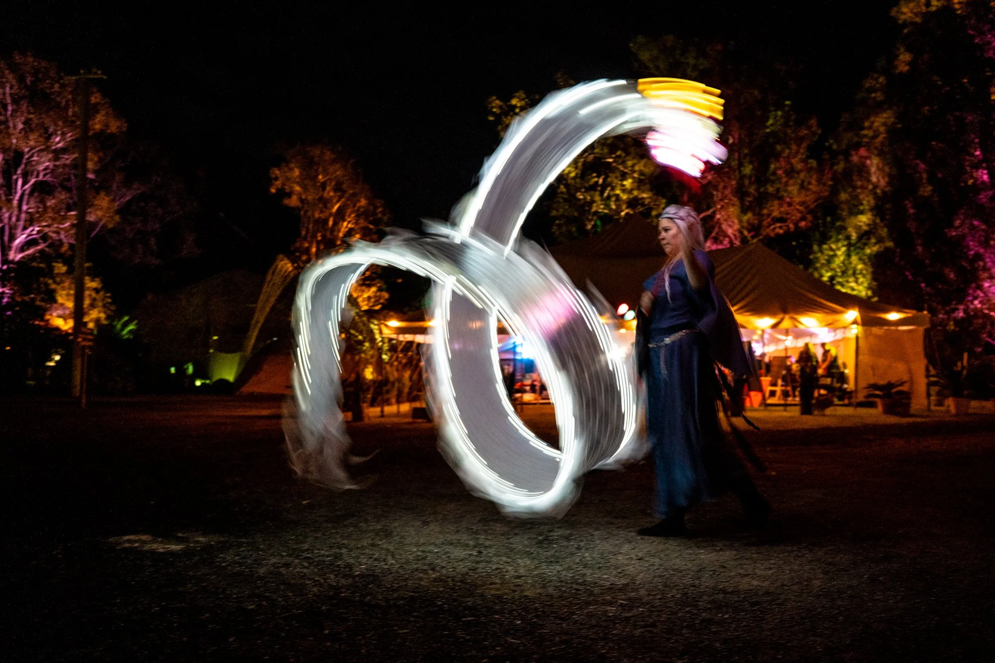 A person dressed as a witch with a hat, performing a light painting with a spinning light object at night, with tents and trees illuminated in the background.