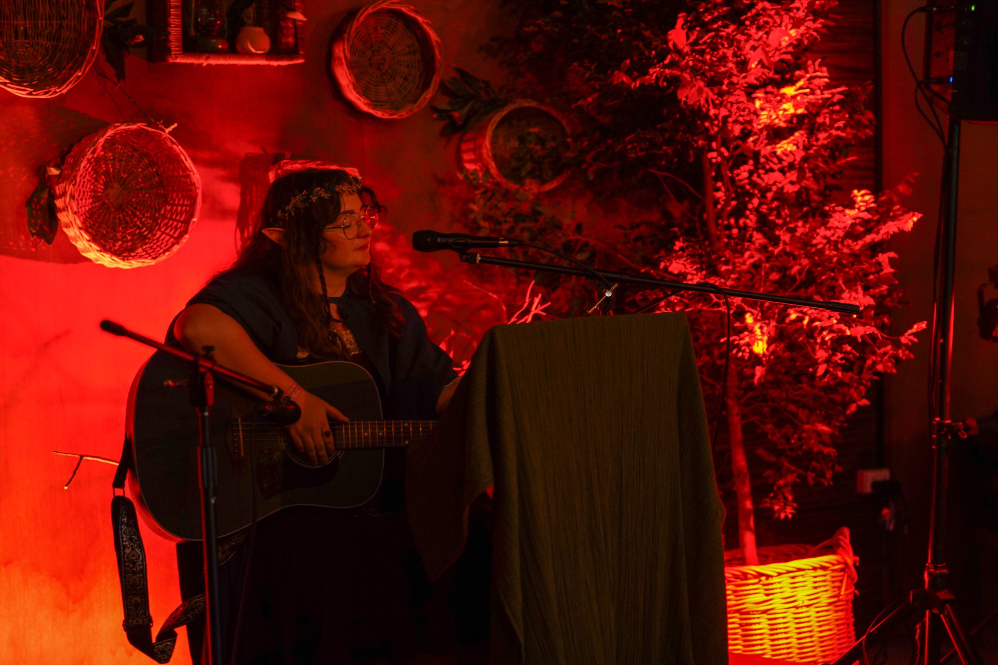 A woman wearing glasses, a floral headband, and elf ears playing an acoustic guitar and singing into a microphone at a cozy indoor venue with red lighting, decorative wicker baskets, and lush greenery.