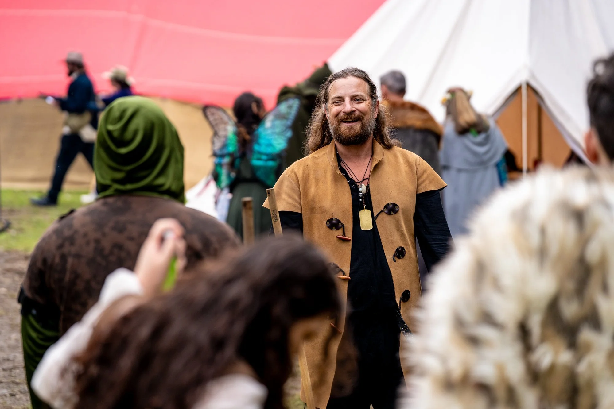 A man with long hair and a beard smiling at a gathering outdoors, with people, tents, and a pinkish-orange sky in the background.