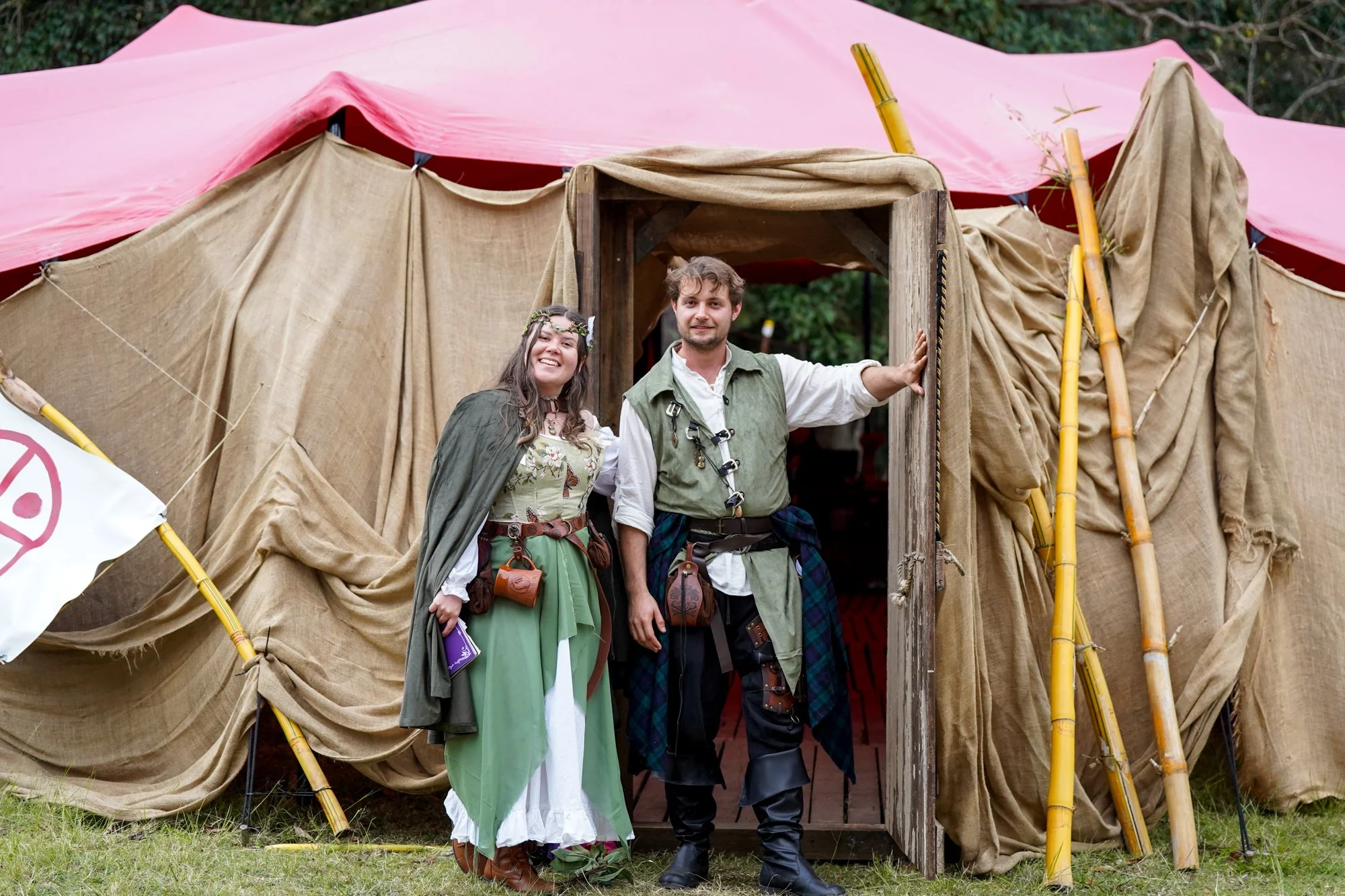 Two people dressed in medieval costumes standing at the entrance of a large beige and pink camping tent with bamboo supports.