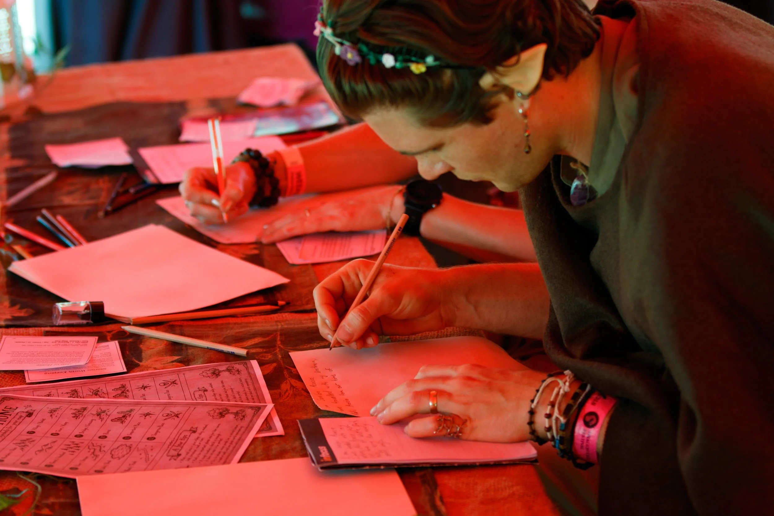 Person writing on paper at a table with pink printed materials, glasses, and pens scattered around.
