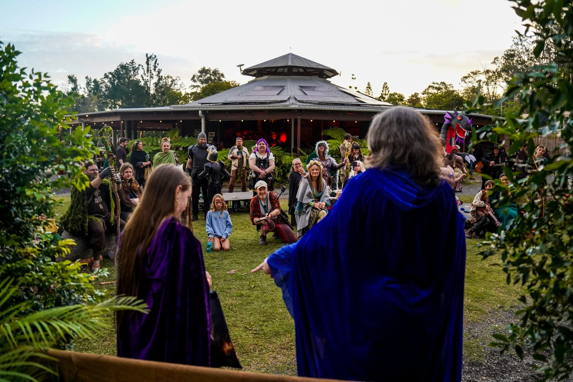 People dressed in medieval and fantasy costumes gather outdoors near a large round building with a roof adorned with solar panels, surrounded by greenery and trees, during what appears to be a themed festival or event.
