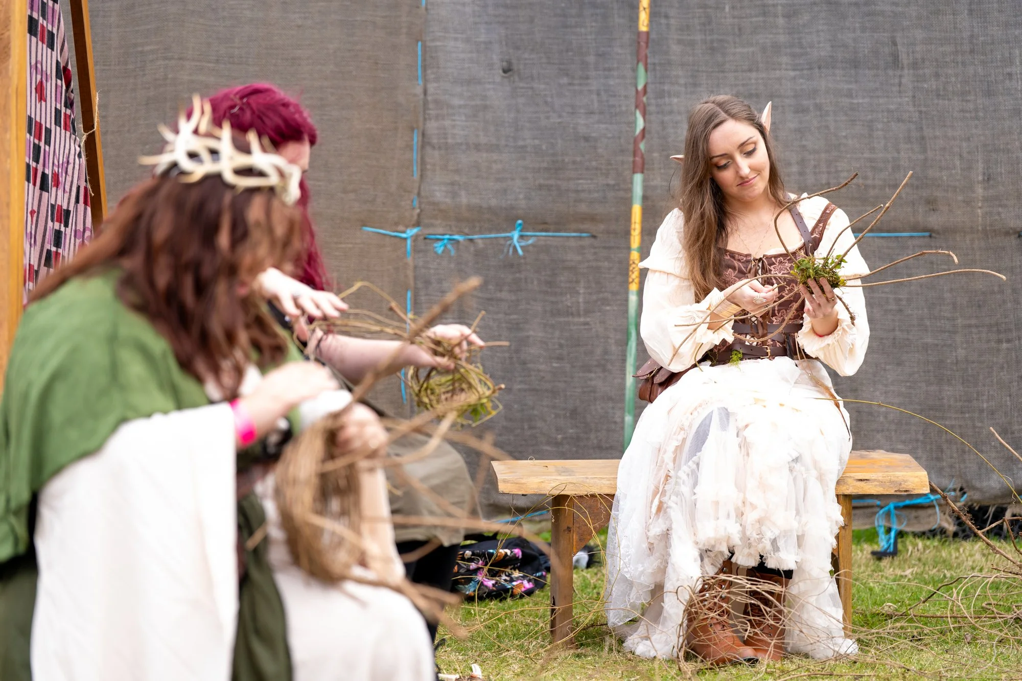Three women sitting on a wooden bench, engaged in a craft activity involving twigs and branches, with a woman in a fantasy or fairy costume with elf ears and a flowing white dress observing or participating, against a background of dark fabric and co
