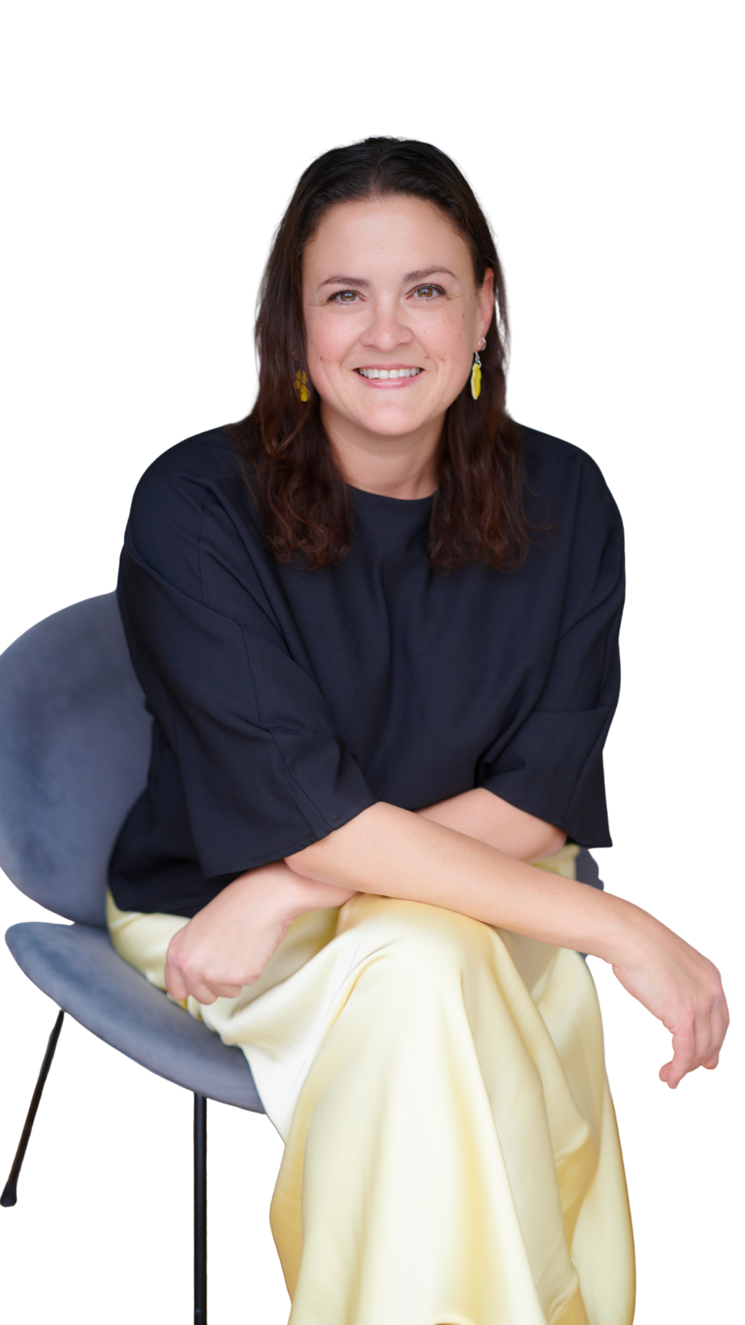 A woman with shoulder-length dark hair, light skin, wearing a black top, yellow earrings, and yellow pants, sitting on a gray chair against a white background, smiling with arms crossed.
