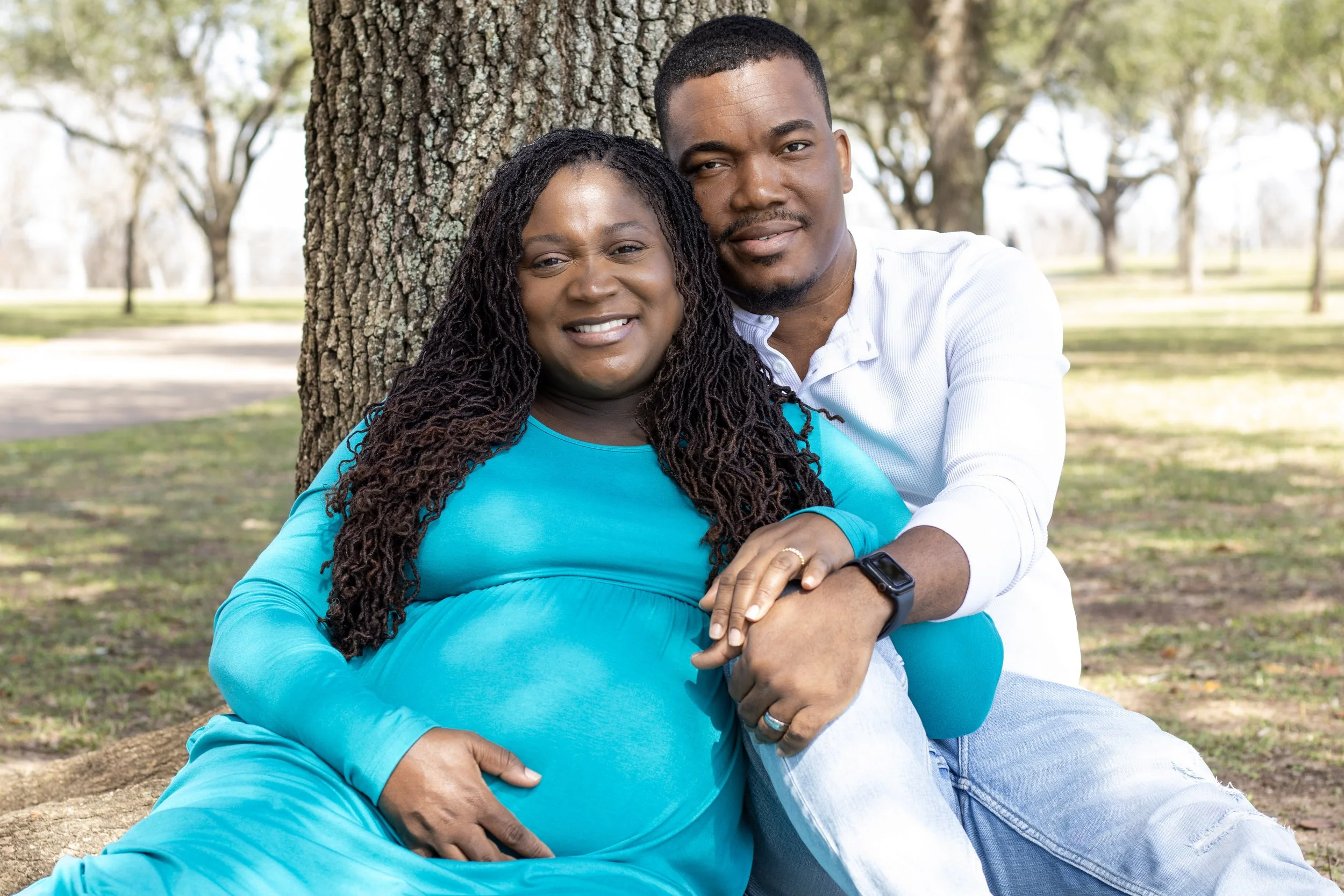 A happy pregnant woman in a blue dress sitting on the ground by a tree, with a man in a white shirt sitting behind her, outdoors in a park.