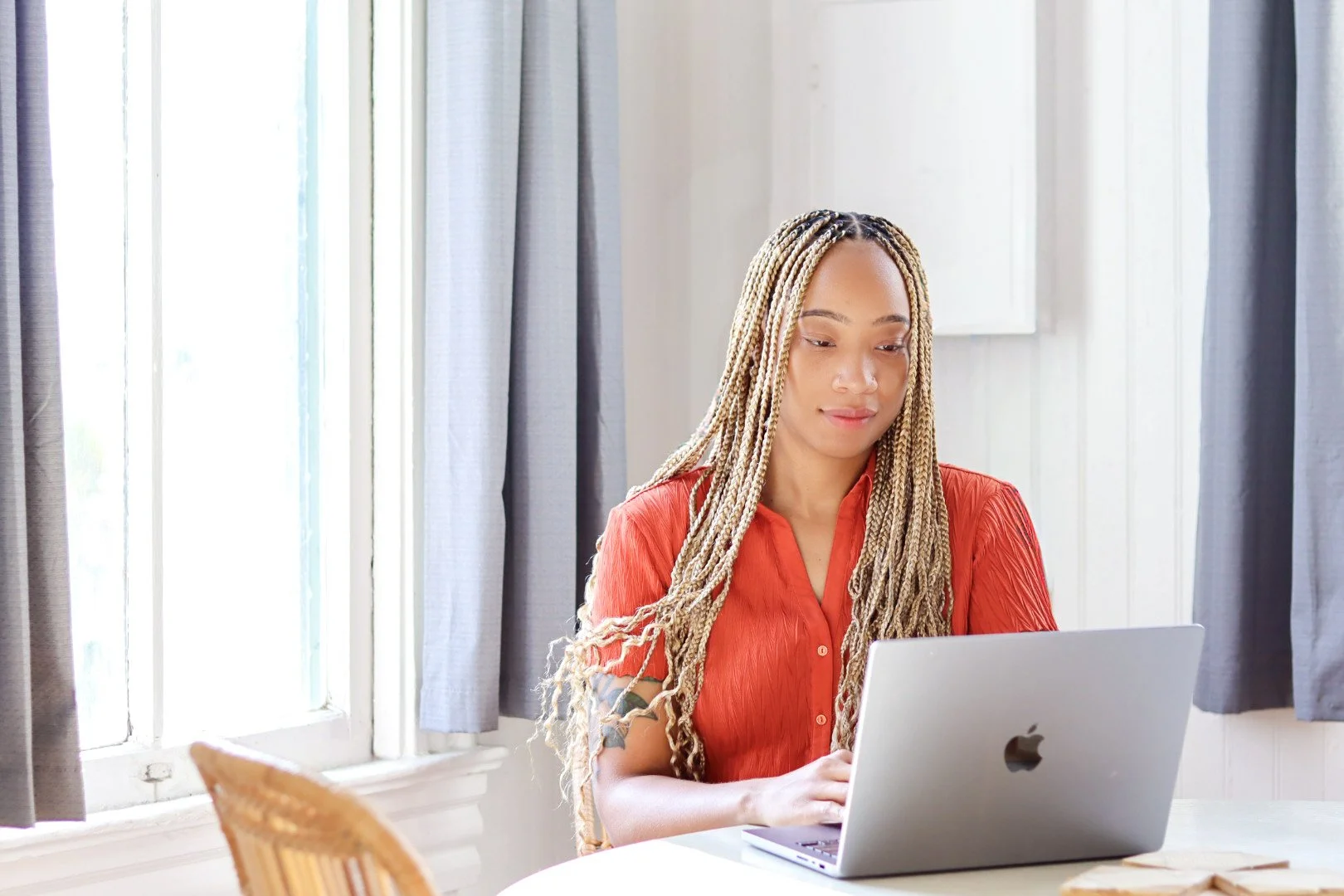 A woman with blonde braided hair wearing a red shirt, sitting at a table, looking at her laptop in a bright room with large windows and gray curtains.