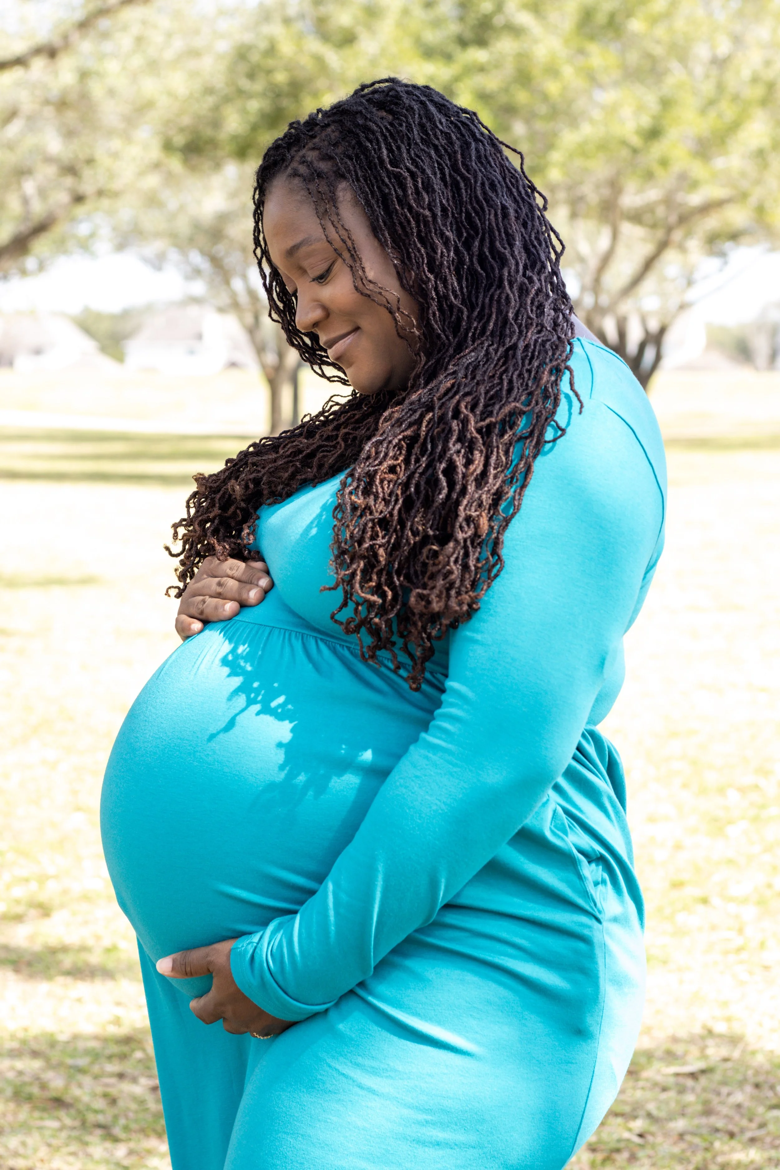 Pregnant woman with long curly hair standing outdoors in a park, wearing a blue dress, smiling gently and looking down at her belly.